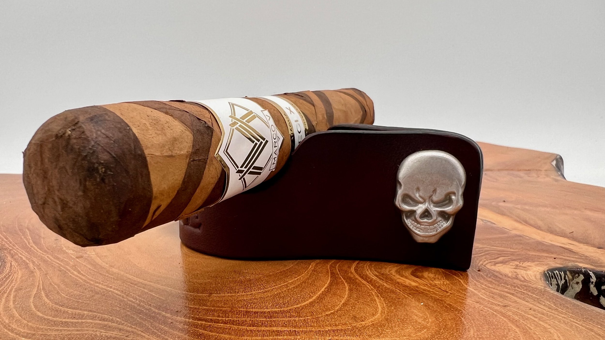 A cigar with a white and gold band resting on a black base with a metallic skull emblem, all placed on a polished wooden surface.