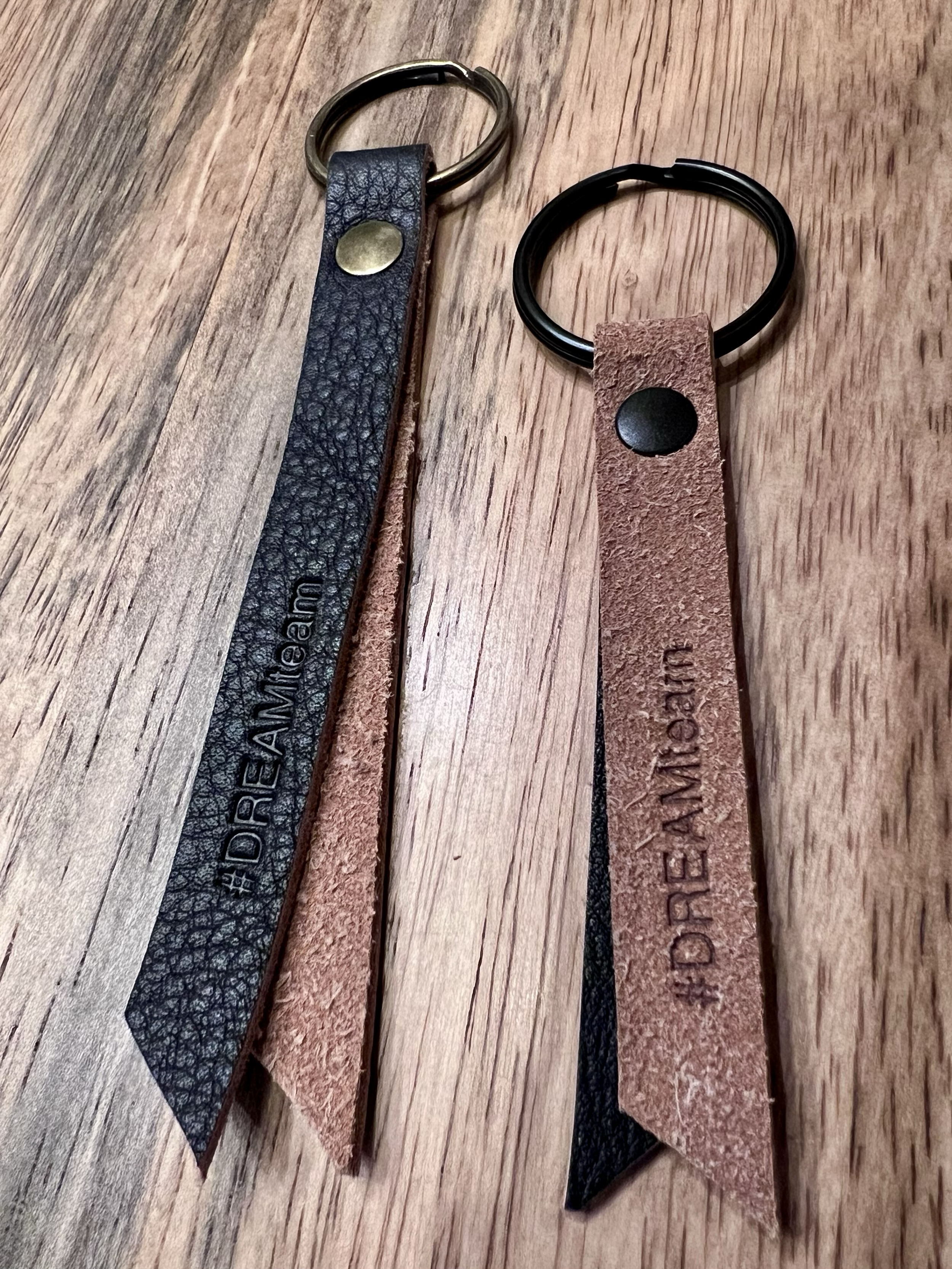 Two leather keychains, one black and one brown, each with a metal ring and a leather strap, with the words 'DREAMTEAM' and 'DREAMPAY' embossed on the straps, placed on a wooden surface.