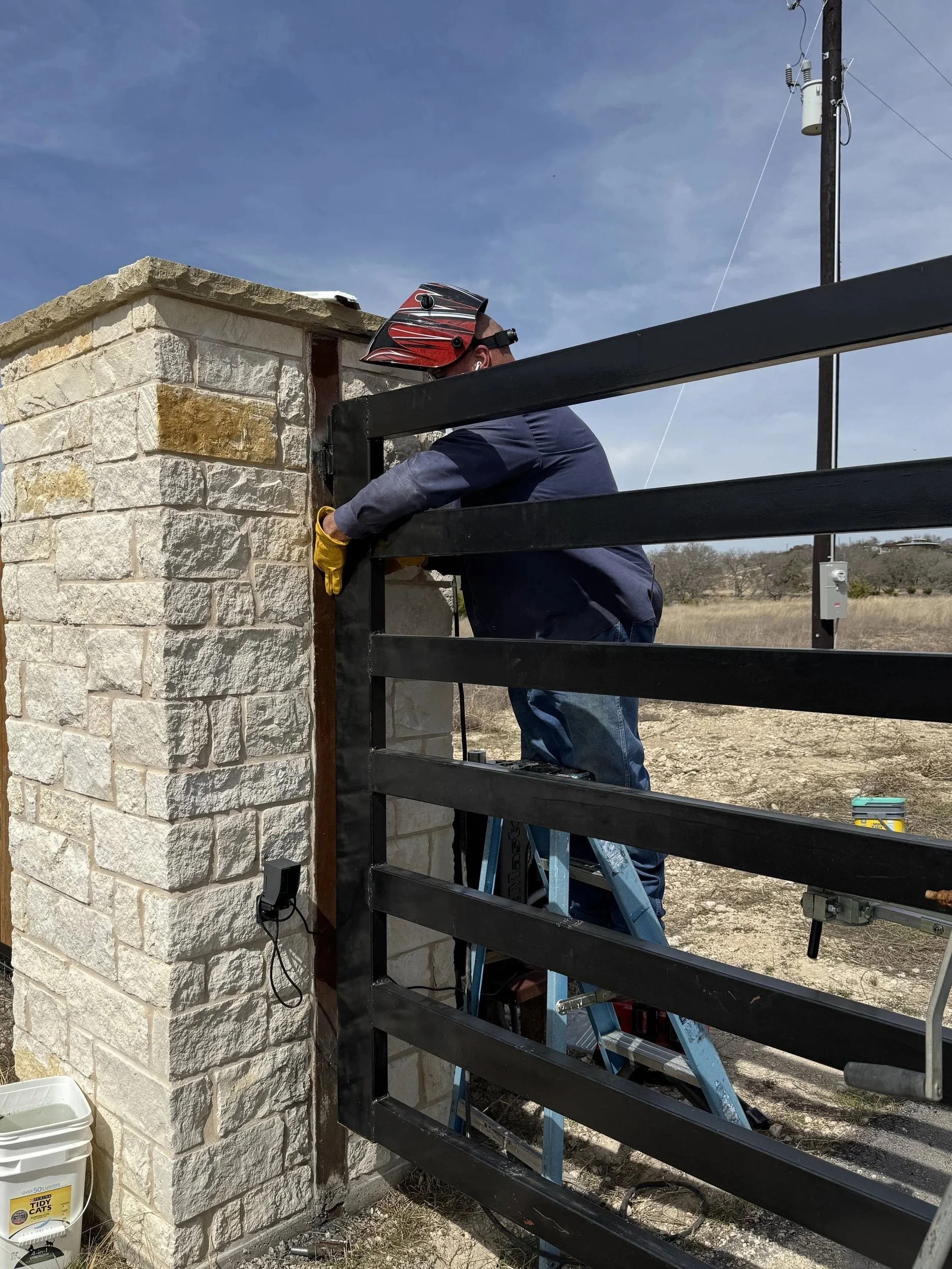 A person wearing a red helmet and yellow gloves is working on installing or repairing a black metal gate on a stone wall, standing on a ladder outdoors with a rural landscape and electrical pole in the background.