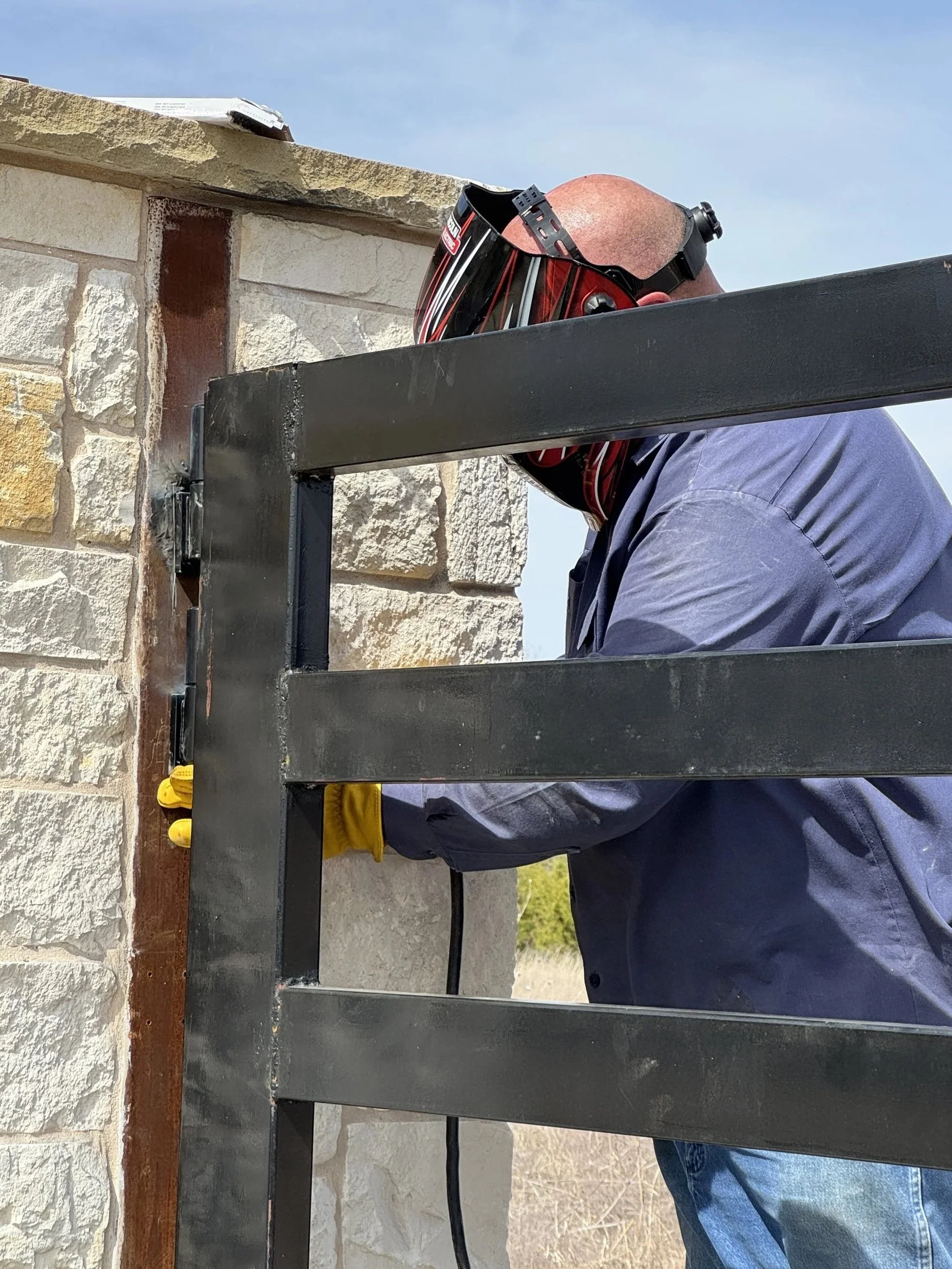 Worker with protective helmet and gloves installing or repairing a metal gate near a stone wall in Gatesville, TX.