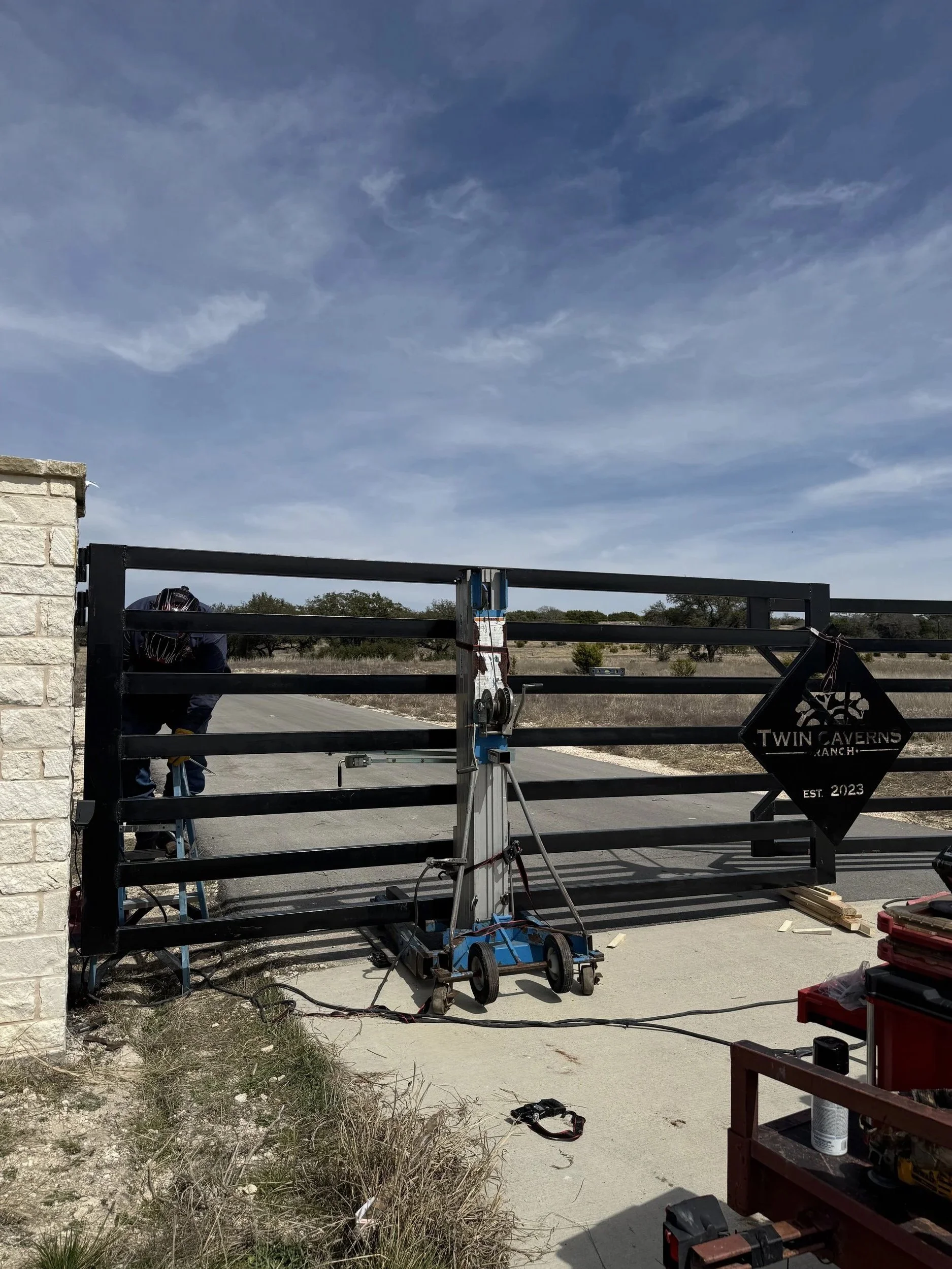 Metal gate with Twin Caverns Ranch sign, a worker on the left, construction tools, and equipment outdoors under a blue sky. After photo of gate project.