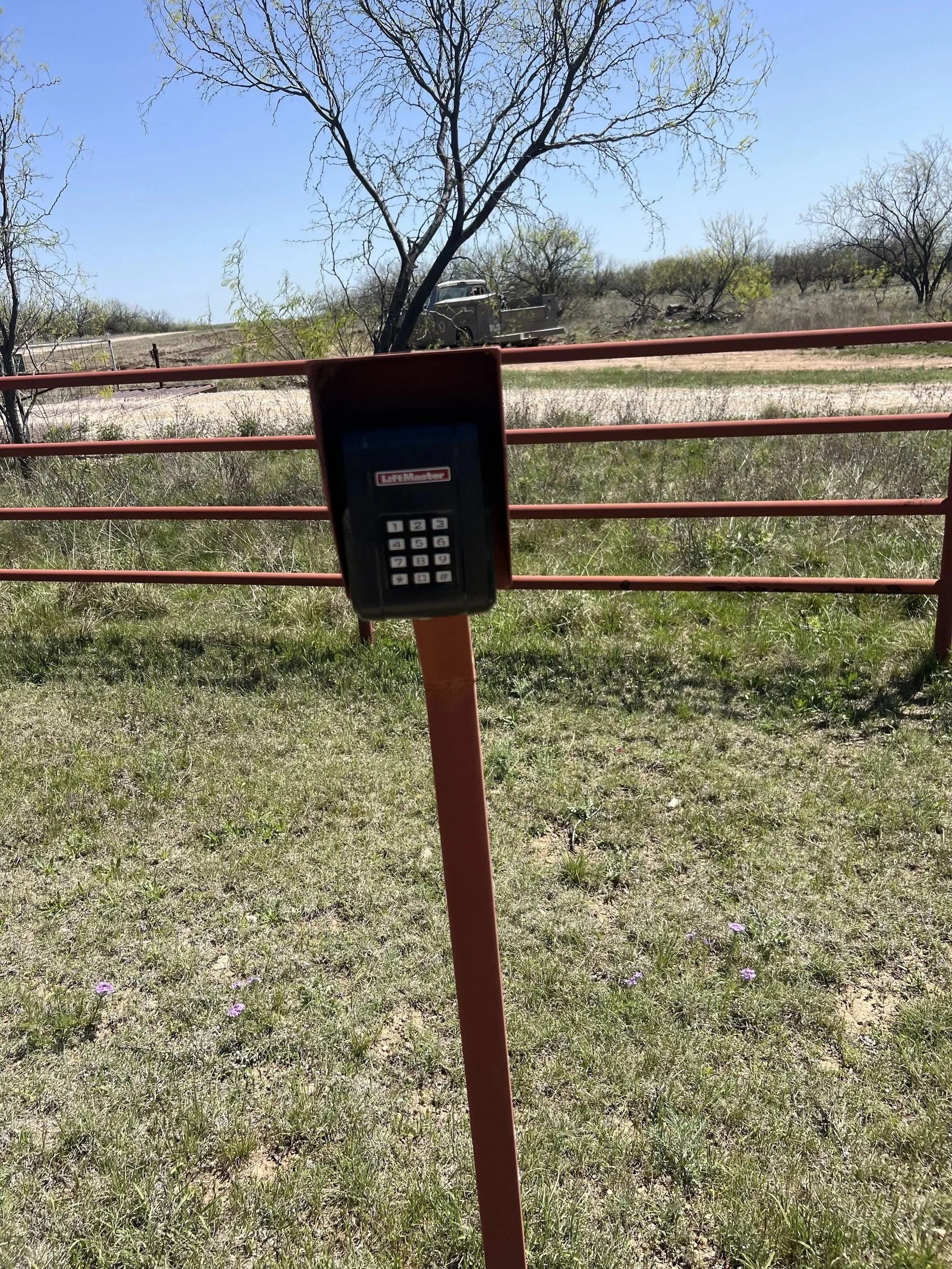 An outdoor scene with a metal gate and an electronic keypad access control mounted on a post. Behind the gate, there is a dirt road, a retro truck, and leafless trees under a clear blue sky.