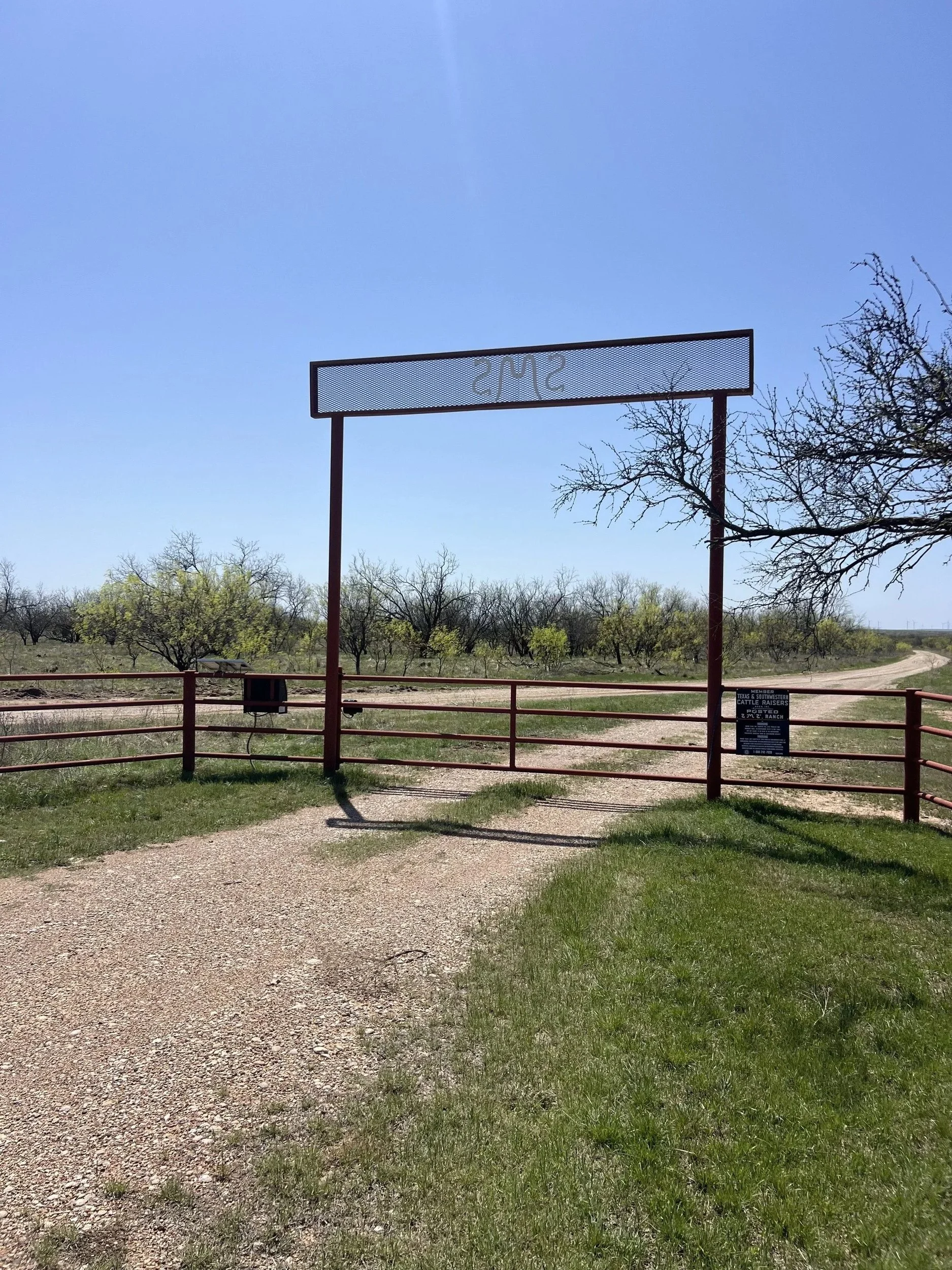 Gate leading to a dirt trail surrounded by grass and trees in a rural area under a clear blue sky.