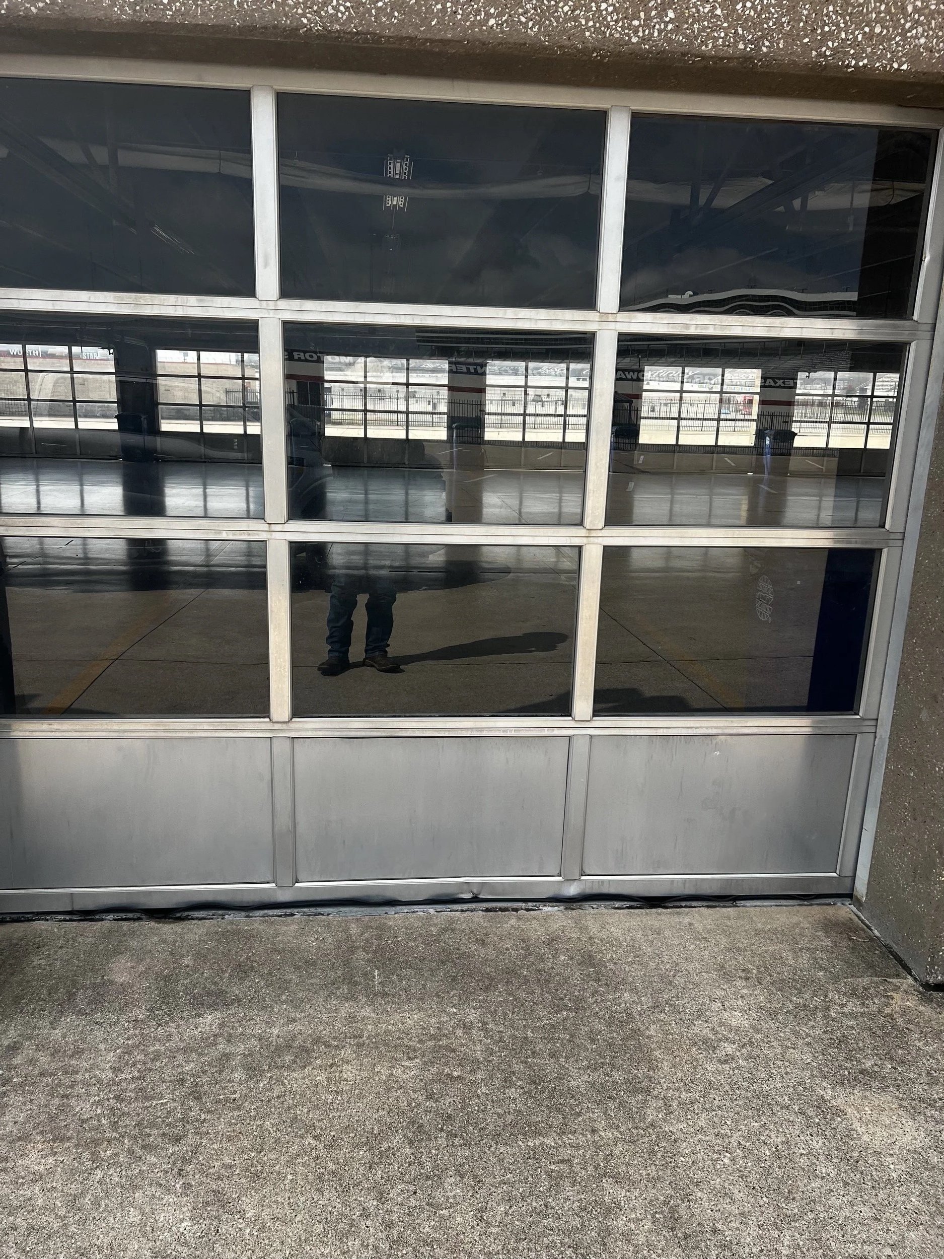 Reflection of a person taking a photo in a large glass garage door with multiple panes; mostly outdoors with some indoor reflections.