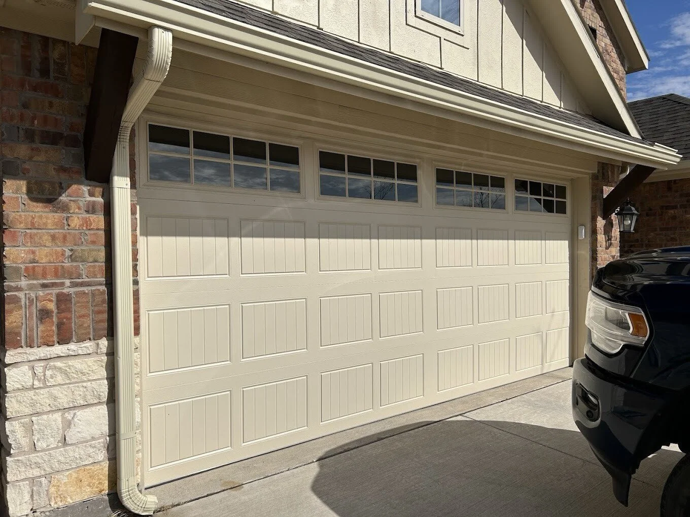 A beige garage door with four small window panes at the top, surrounding brick and siding house, driveway, and part of a black vehicle visible.