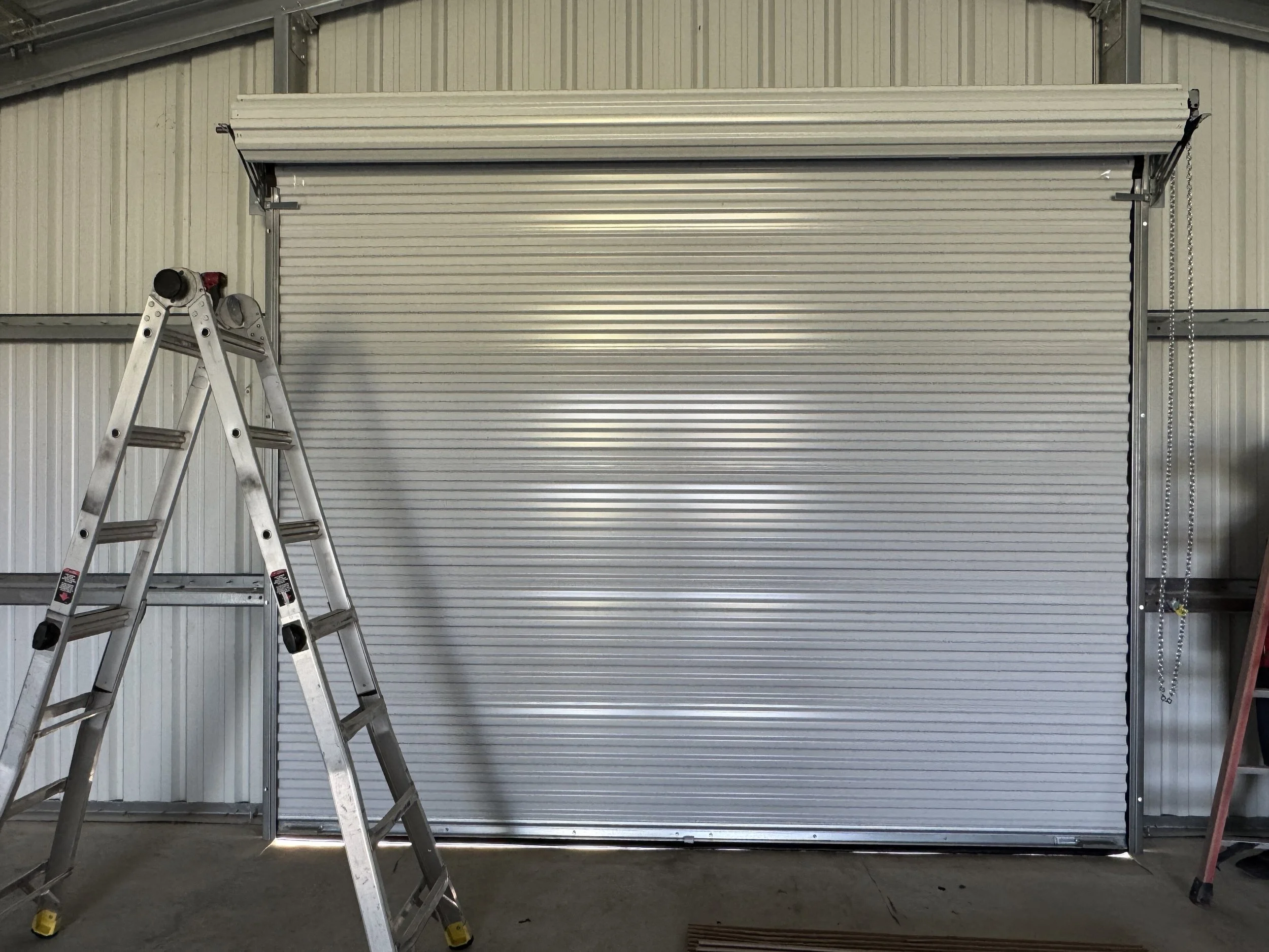 A closed metal roll-up garage door in an industrial setting, flanked by ladders and building materials.