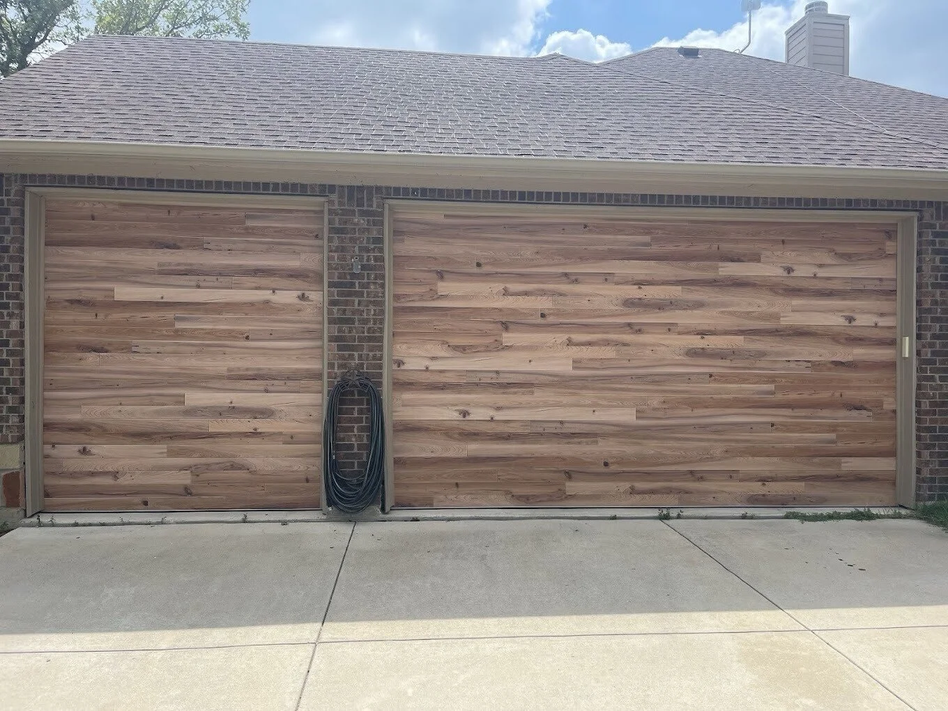 Two wooden garage doors with a brick wall in between, a coiled garden hose hanging on the wall, and a concrete driveway in front.