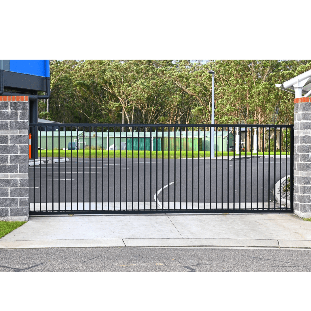 Black metal gate between brick pillars, opening into a parking lot with trees in the background.