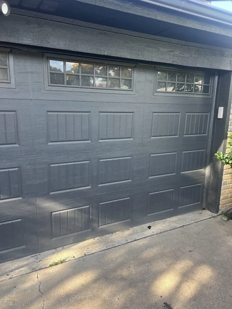 A black garage door with rectangular panels and two small windows at the top, set in a suburban home with a concrete driveway.