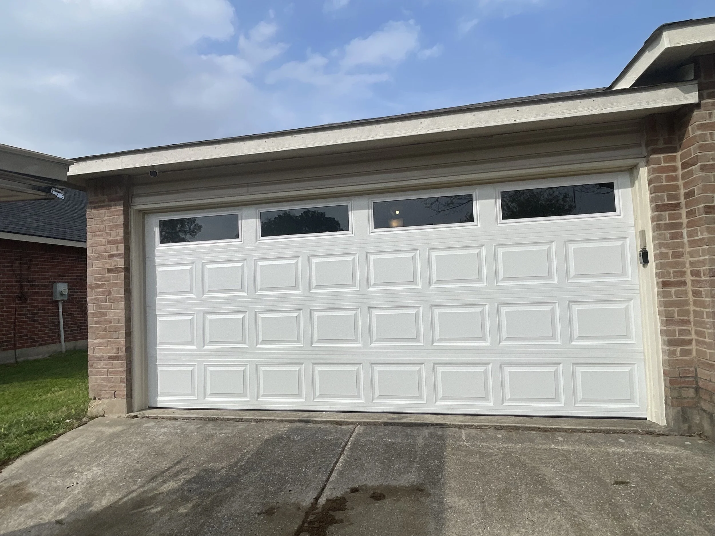 A white garage door with rectangular panels and three horizontal windows at the top, set in a brick garage structure with a sloped roof and a concrete driveway in front.