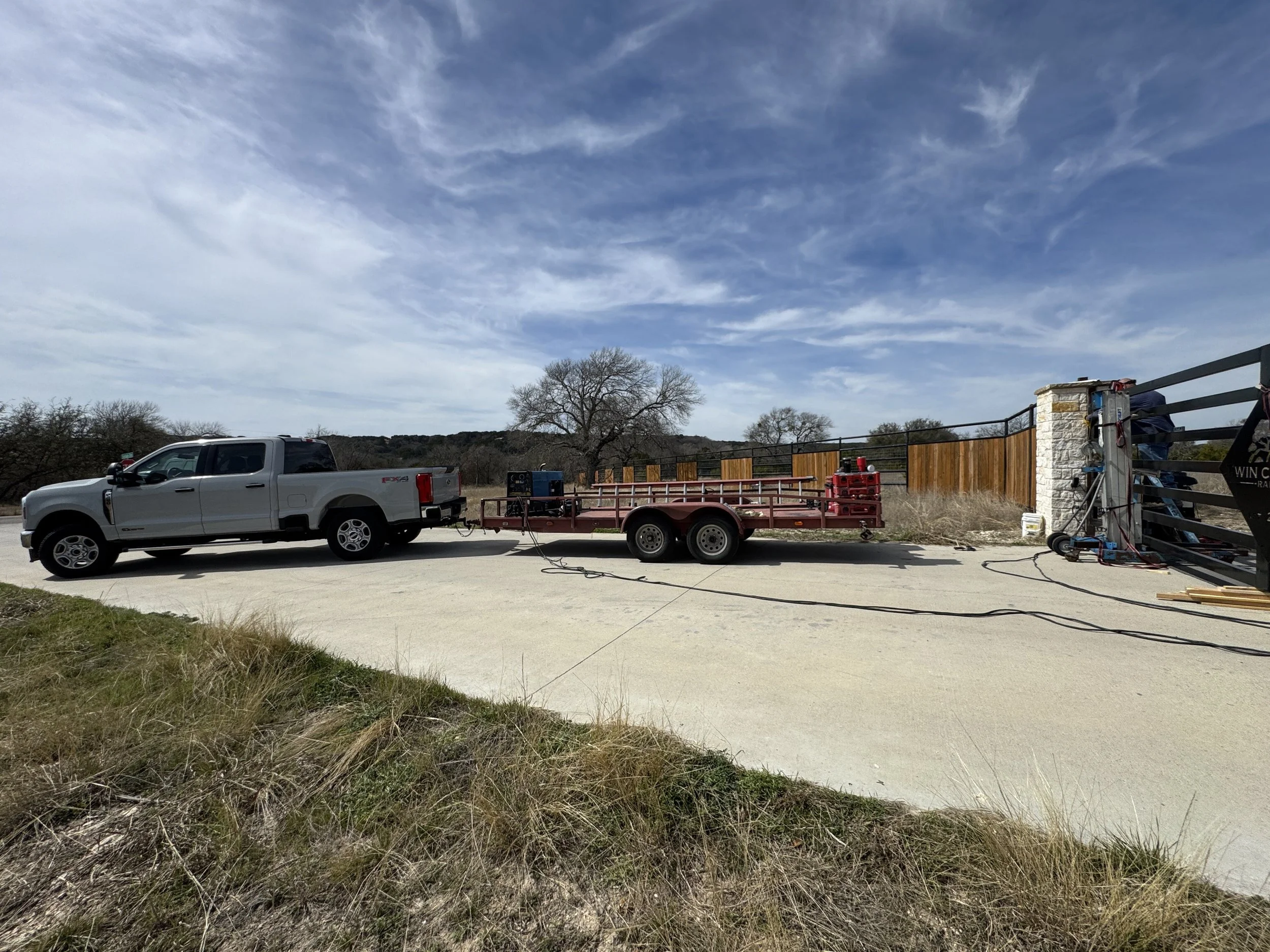 A gray pickup truck towing a trailer parked on a concrete driveway under a partly cloudy sky, with a fence and trees in the background.