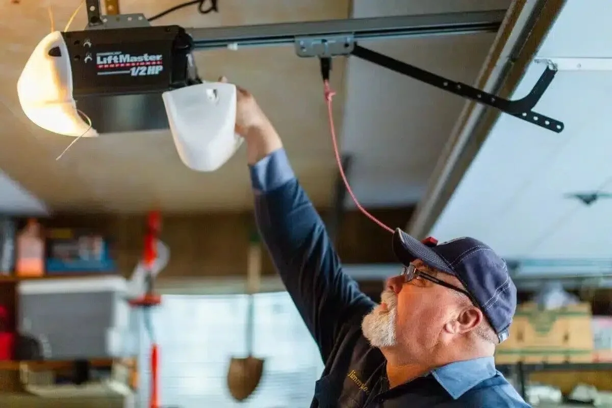 An older man with a gray beard and glasses, wearing a baseball cap and a dark shirt, is reaching up to the garage door opener to activate it.
