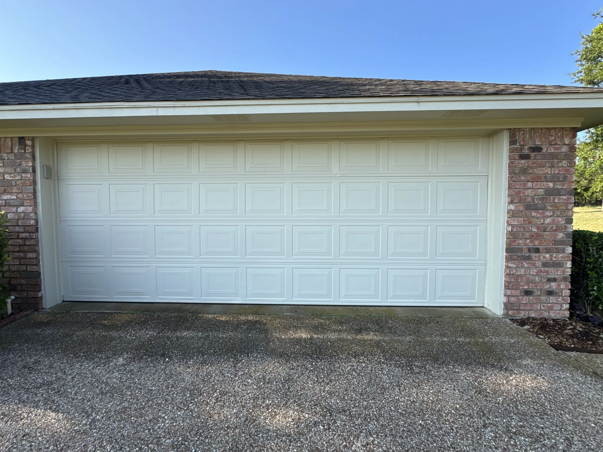 White garage door with brick sides and a shingled roof beneath a clear blue sky.