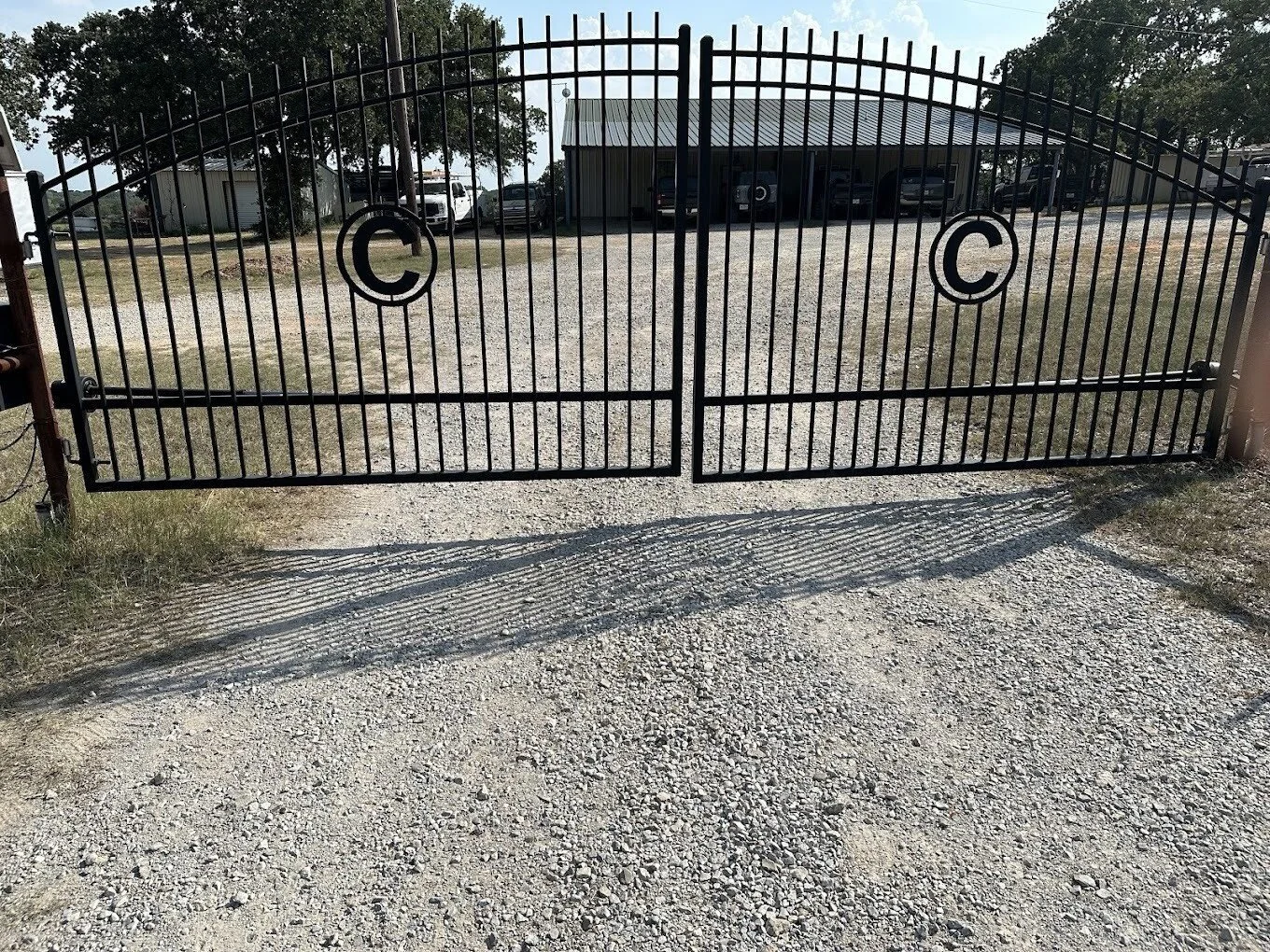 A black metal double gate with decorative circles on each side, casting shadows on a gravel driveway. Behind the gate, there are several parked cars and metal buildings with trees in the background.