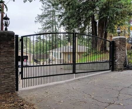 A black metal gate with decorative top, flanked by stone pillars, closing a driveway in a residential area with trees and houses in the background.