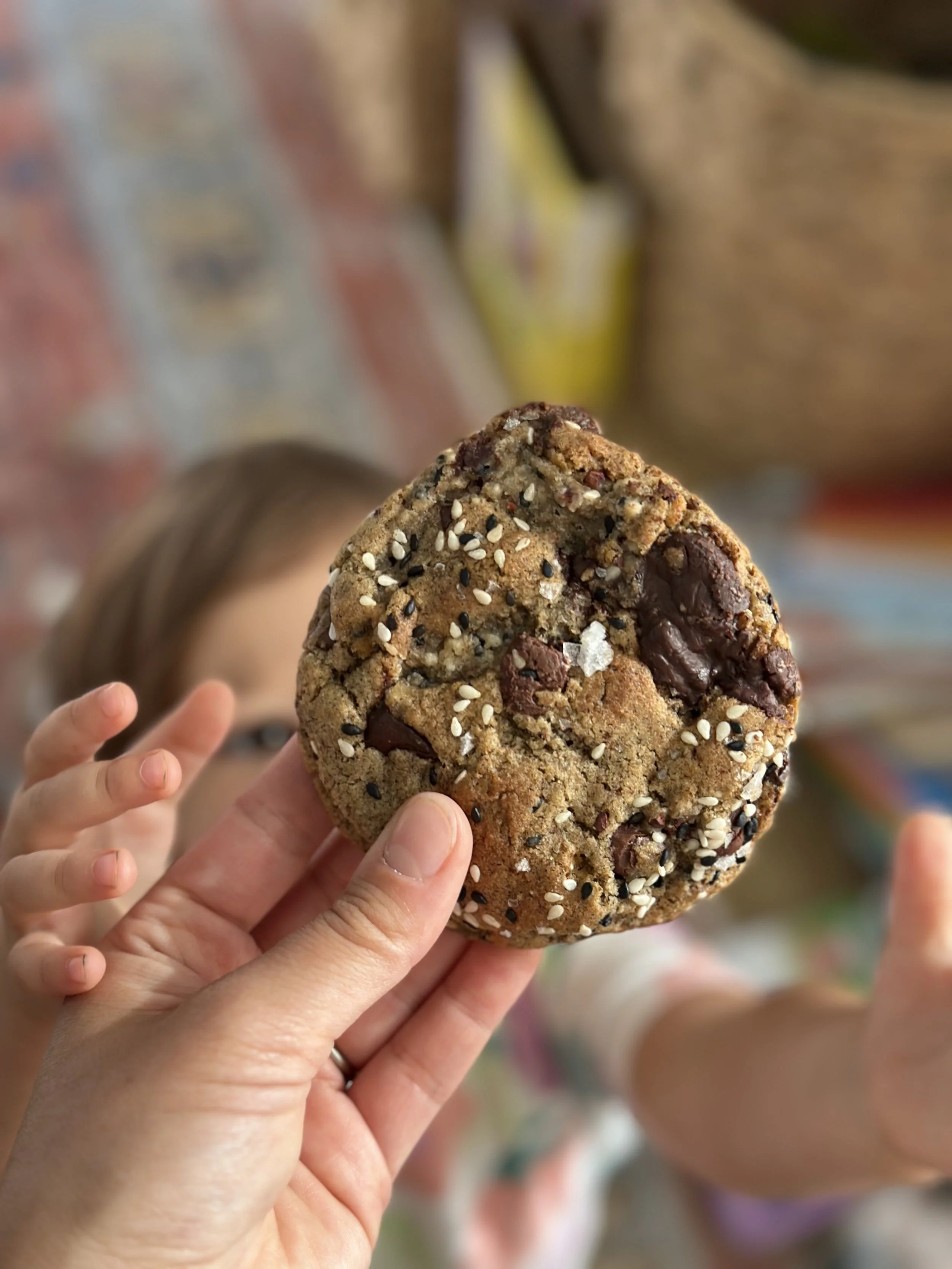 Close-up of a large, homemade chocolate chip cookie with sesame by Chicky's Sourdough, held by an adult hand, with a young child's hand reaching up to it - a little sweet treat for the whole family delivered right to your home.