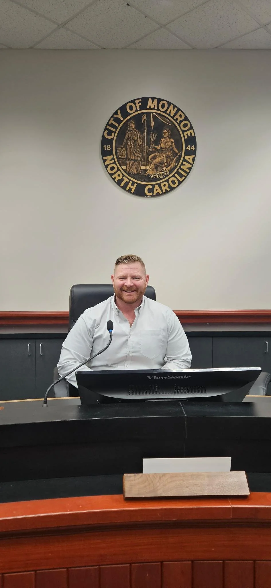 A man with red hair and a beard sitting at a desk in front of a computer, smiling at the camera. Behind him on the wall is a seal of the city of Monroe, North Carolina with the date 1844.