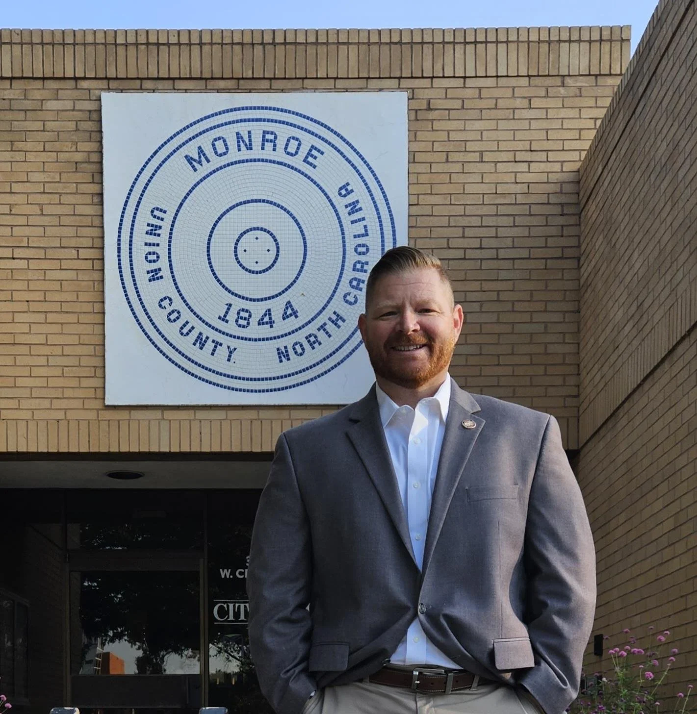 A man in a gray suit and white shirt standing outside a building with a large white sign that reads 'Monroe North County North Bowling 1844'.