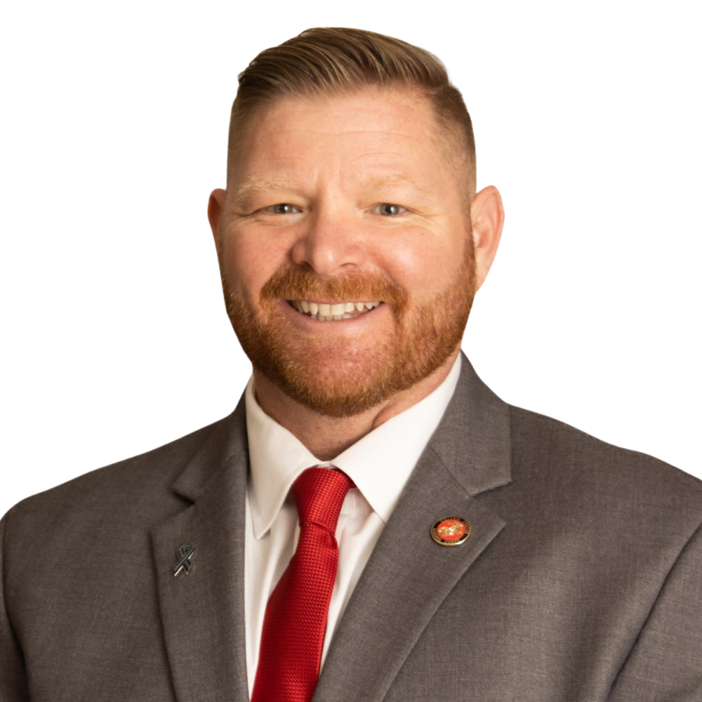 A man with a beard and red hair wearing a grey suit with a white shirt and red tie, smiling.