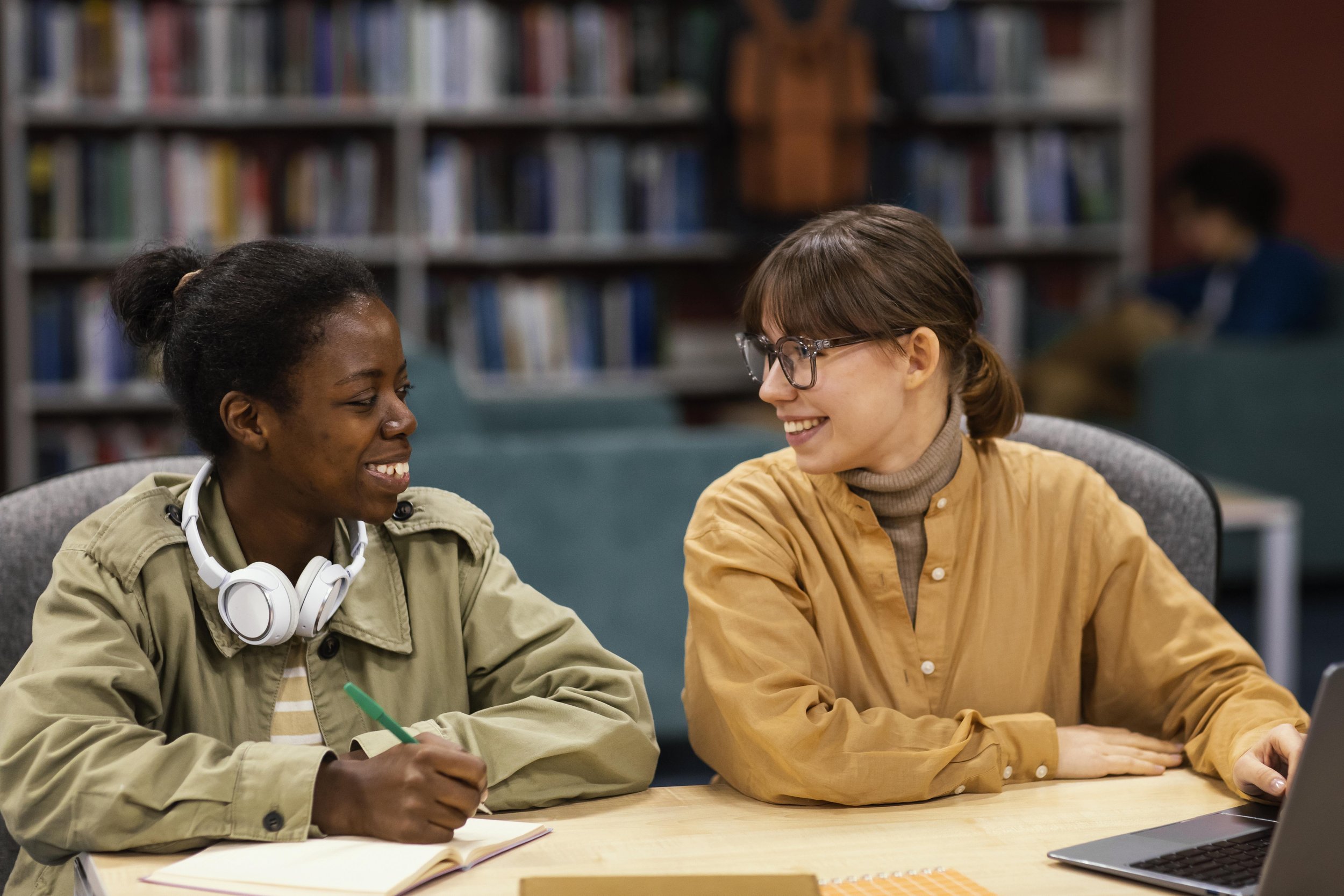 Two smiling women sitting at a table in a library, engaging in conversation. One woman is wearing headphones around her neck and holding a pen, with a notebook in front of her. The other woman is wearing glasses and a tan shirt, with a laptop open in front of her.