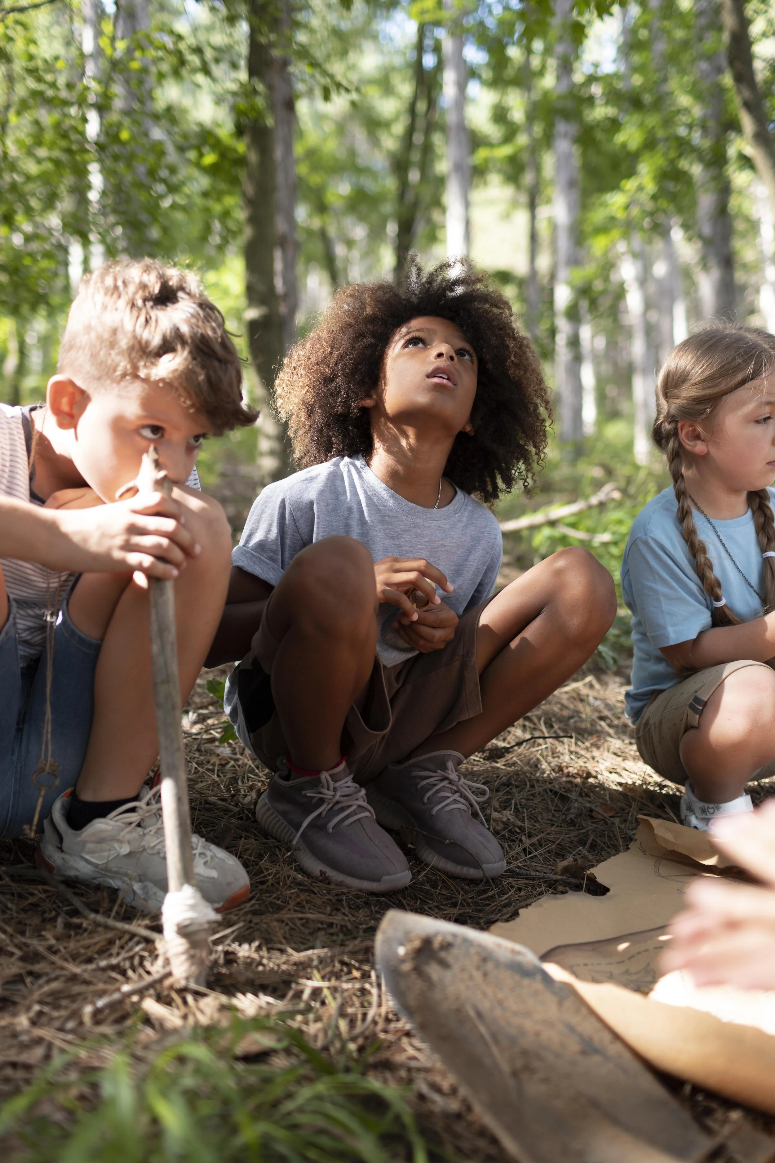 Three children sitting on the ground in a forest, looking upwards with curious expressions, surrounded by trees and sunlight filtering through leaves.