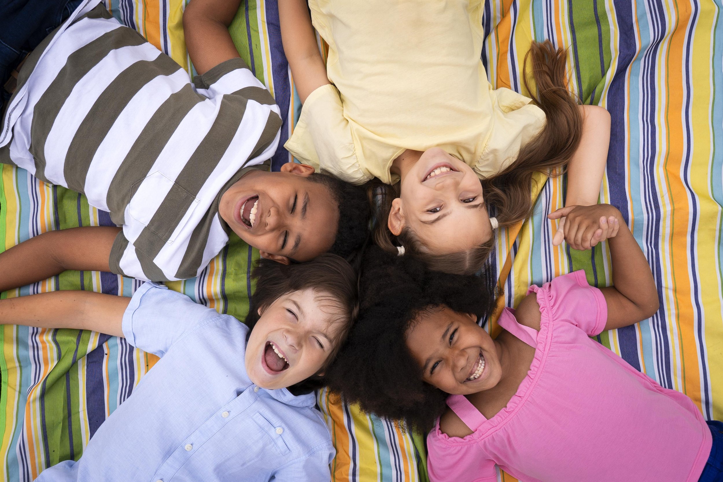 Five children lying on a striped blanket, smiling and laughing with their heads close together.