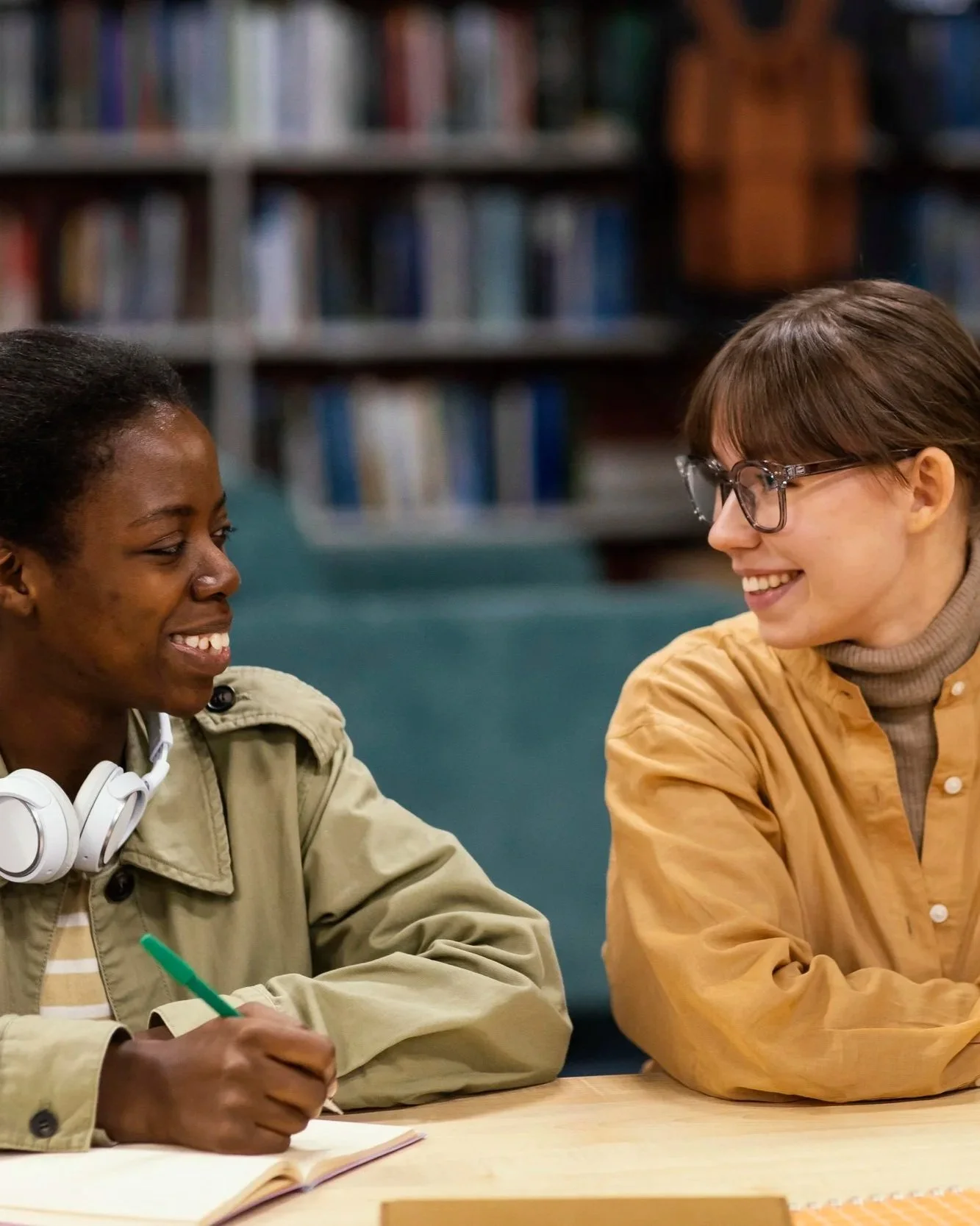 Two young women sitting at a table in a library, smiling and talking to each other, with bookshelves in the background.