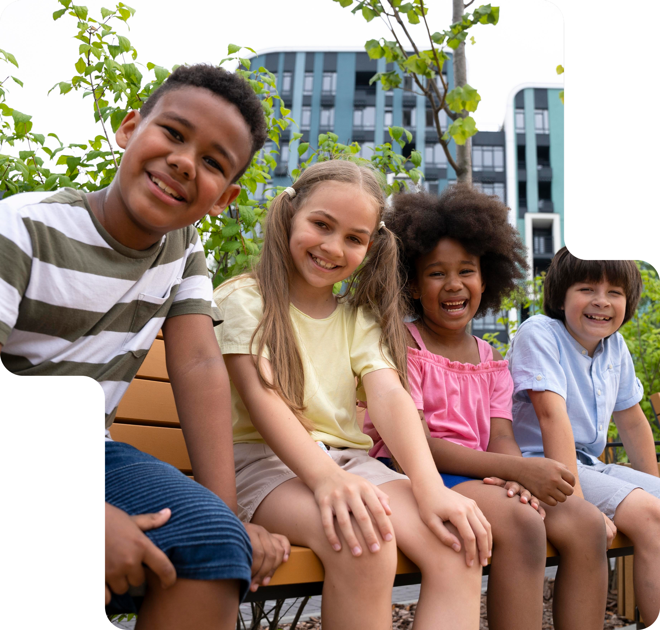 Four children sitting on a park bench, smiling. There are green plants and a modern building in the background.