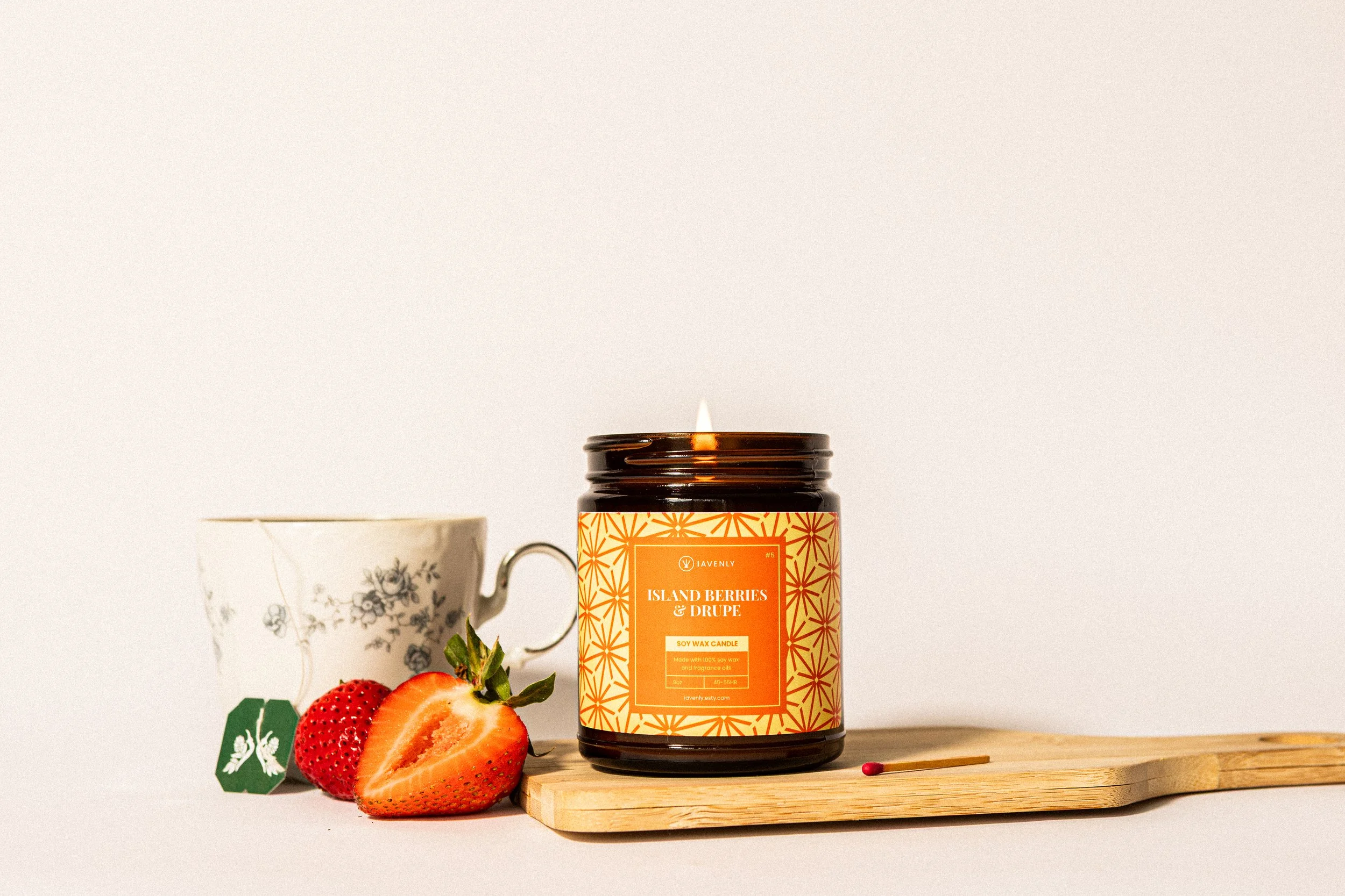 A lit soy wax candle labeled 'Island Berries & Drupe' on a wooden board, with a strawberry half, a strawberry, a tea infuser, and a floral-patterned teacup in the background.