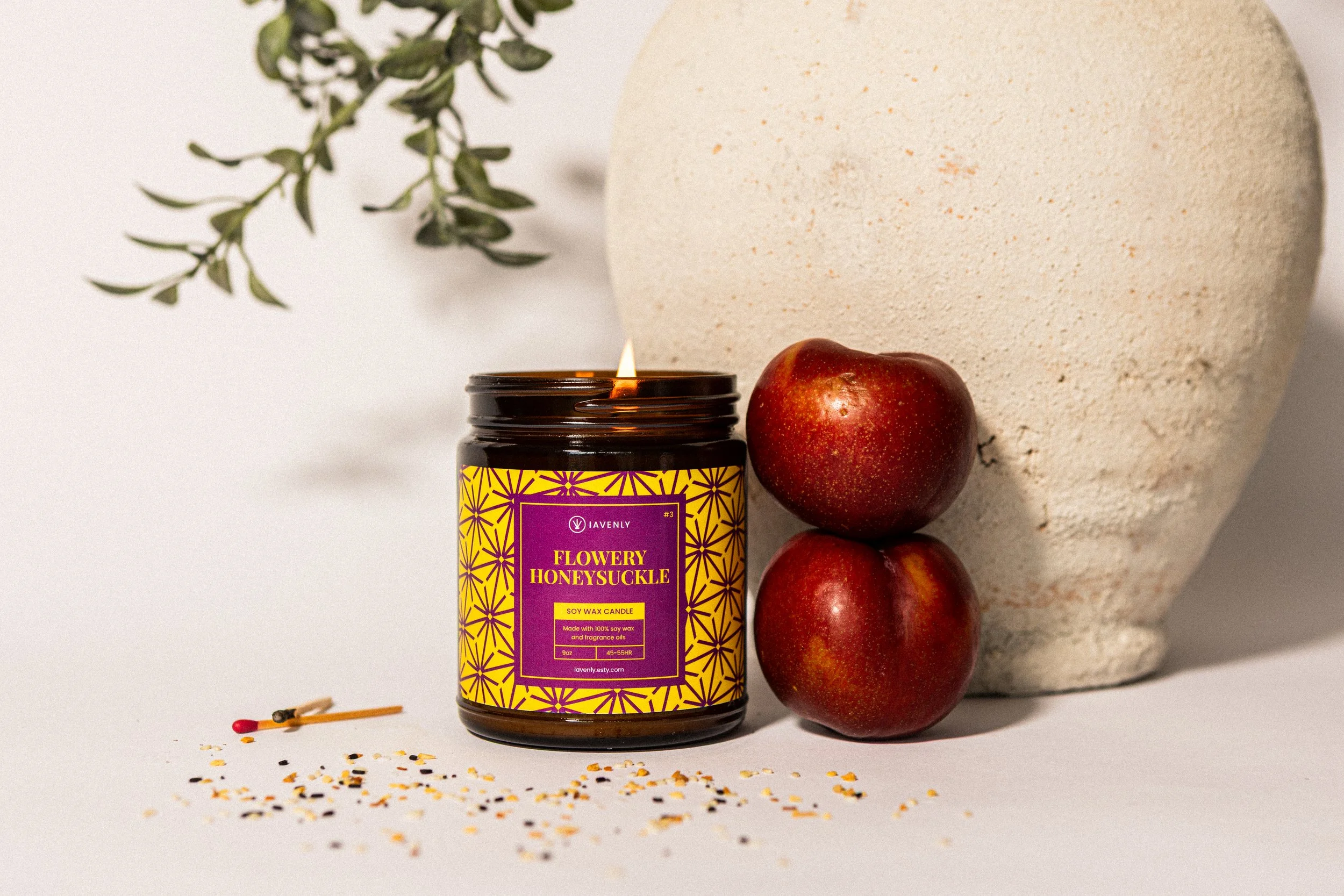 A lit soy wax candle labeled 'Flowery Honeysuckle' with red apples, a large white textured vase, a matchstick near scattered colorful confetti, and a green leafy branch in the background.