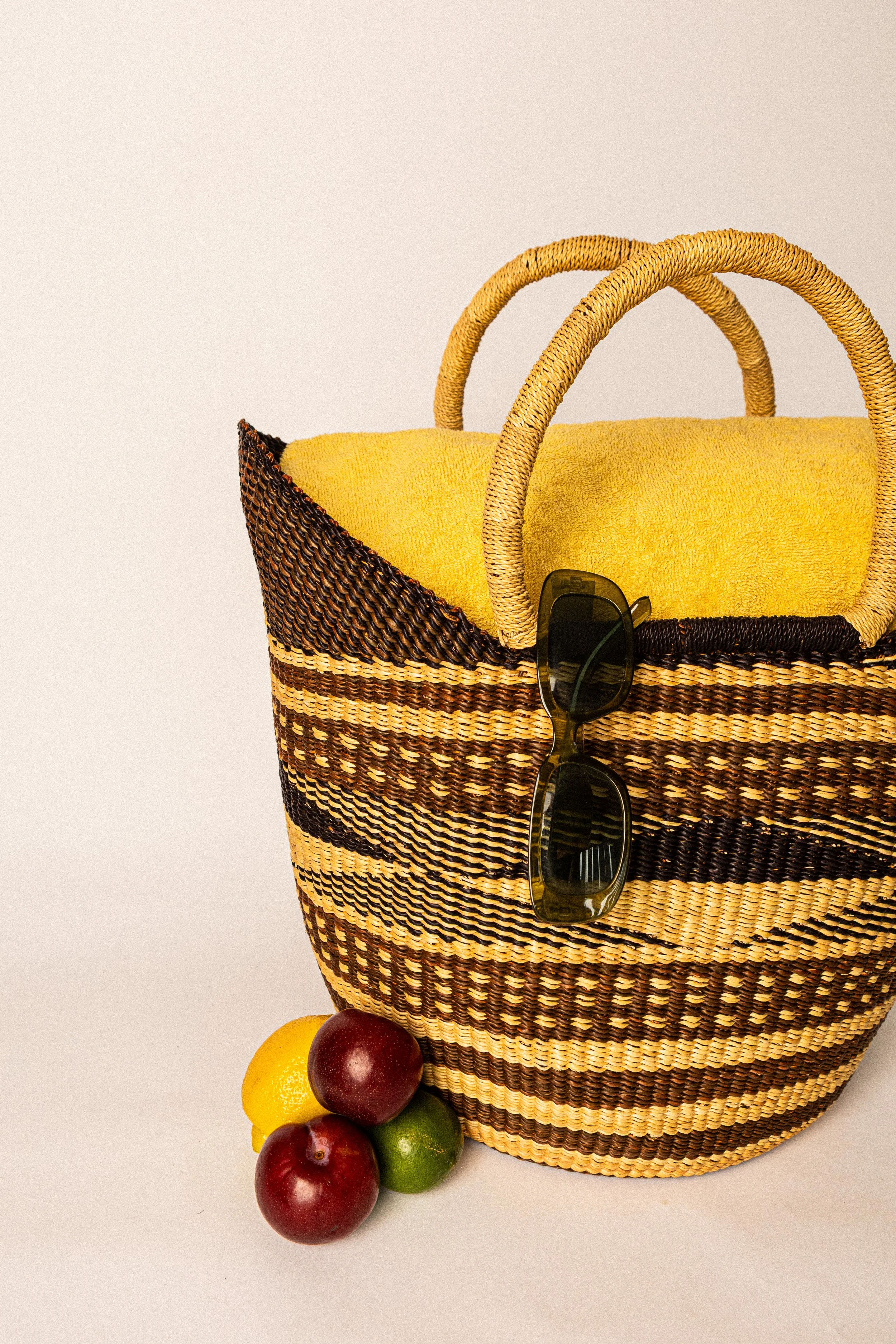 A woven straw beach bag with yellow towel inside, sunglasses hanging from the strap, and three fruits—limes and plums—placed next to it on a white background.