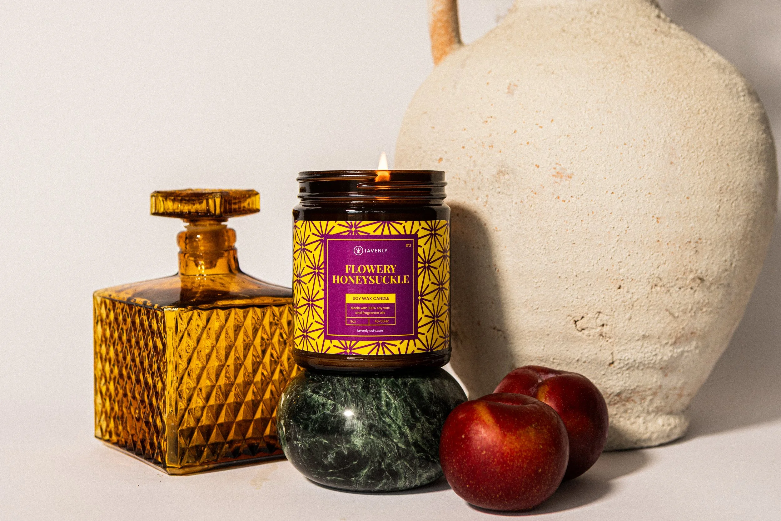 Decorative still life with amber glass bottle, lit candle in a jar labeled "Flowery Honeysuckle" soy wax candle, green marble base, white textured vase, and two red apples.