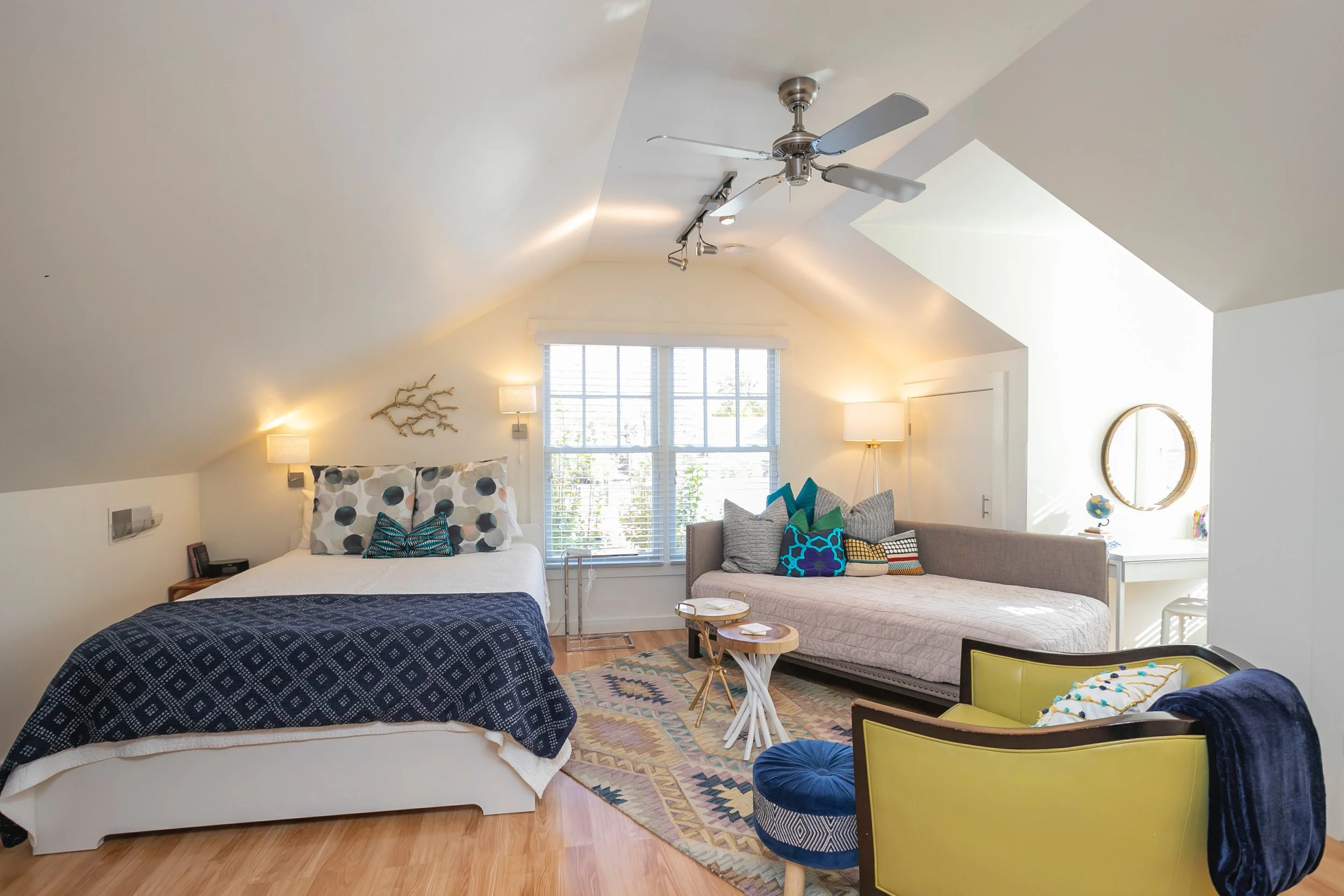 A stylish bedroom with a sloped ceiling, containing a bed with a black and white patterned quilt and pillows, a sitting area with a beige daybed and colorful pillows, a pair of small wooden side tables, a yellow armchair with a pillow and a dark blanket, a window with white blinds, standing lamps, a mirror, and decorative wall accents, illuminated by natural and warm lighting.