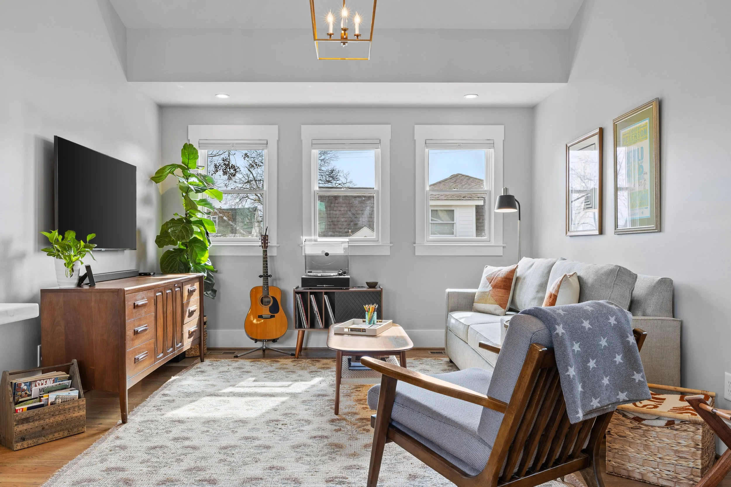 Bright living room with a white sofa, striped blue rug, wooden coffee table, gray armchair, wall-mounted TV, three windows, and a decorative light fixture.