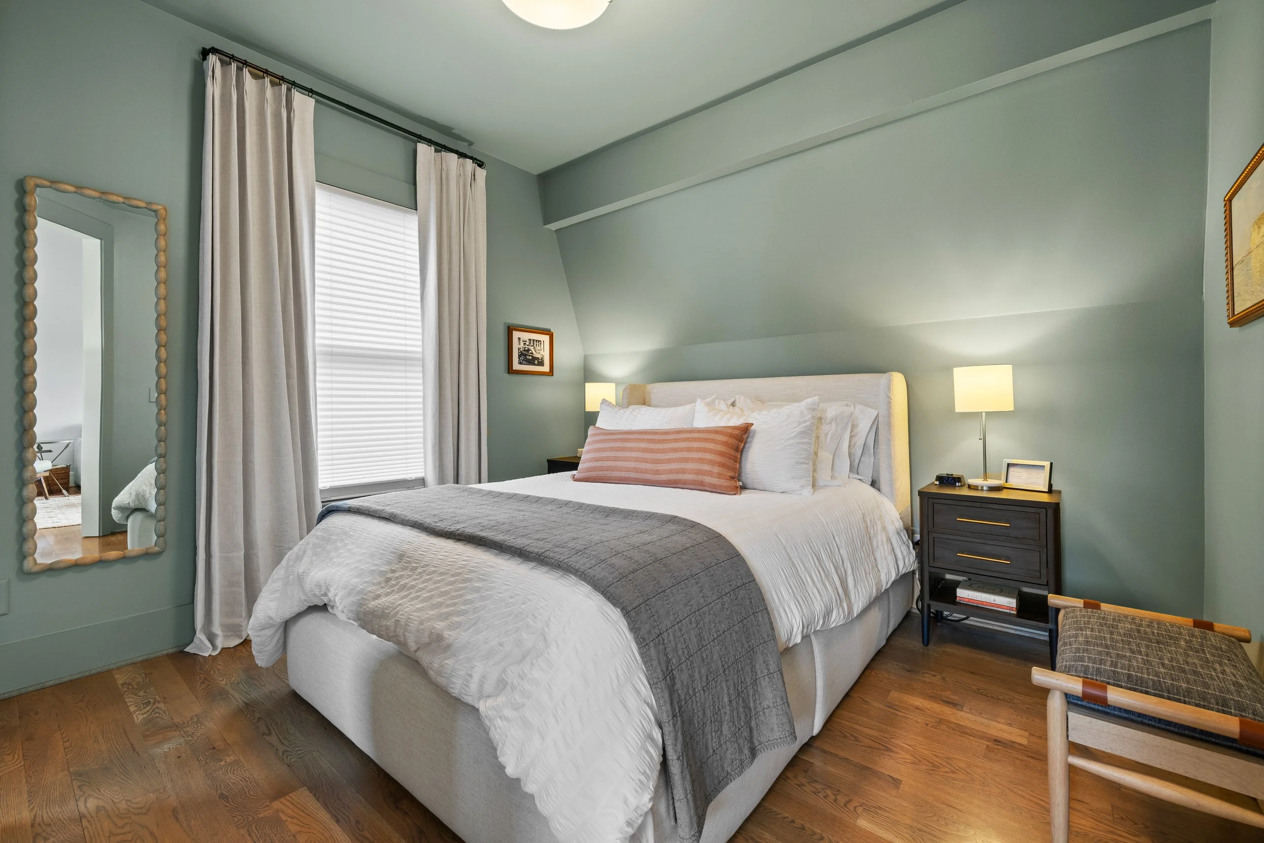 A neatly made bed with white linens, decorative pillows, and a gray throw blanket in a bright bedroom. There are nightstands with lamps on either side of the bed, a mirror reflecting part of the room, and a large window with patterned curtains allowing natural light to enter.