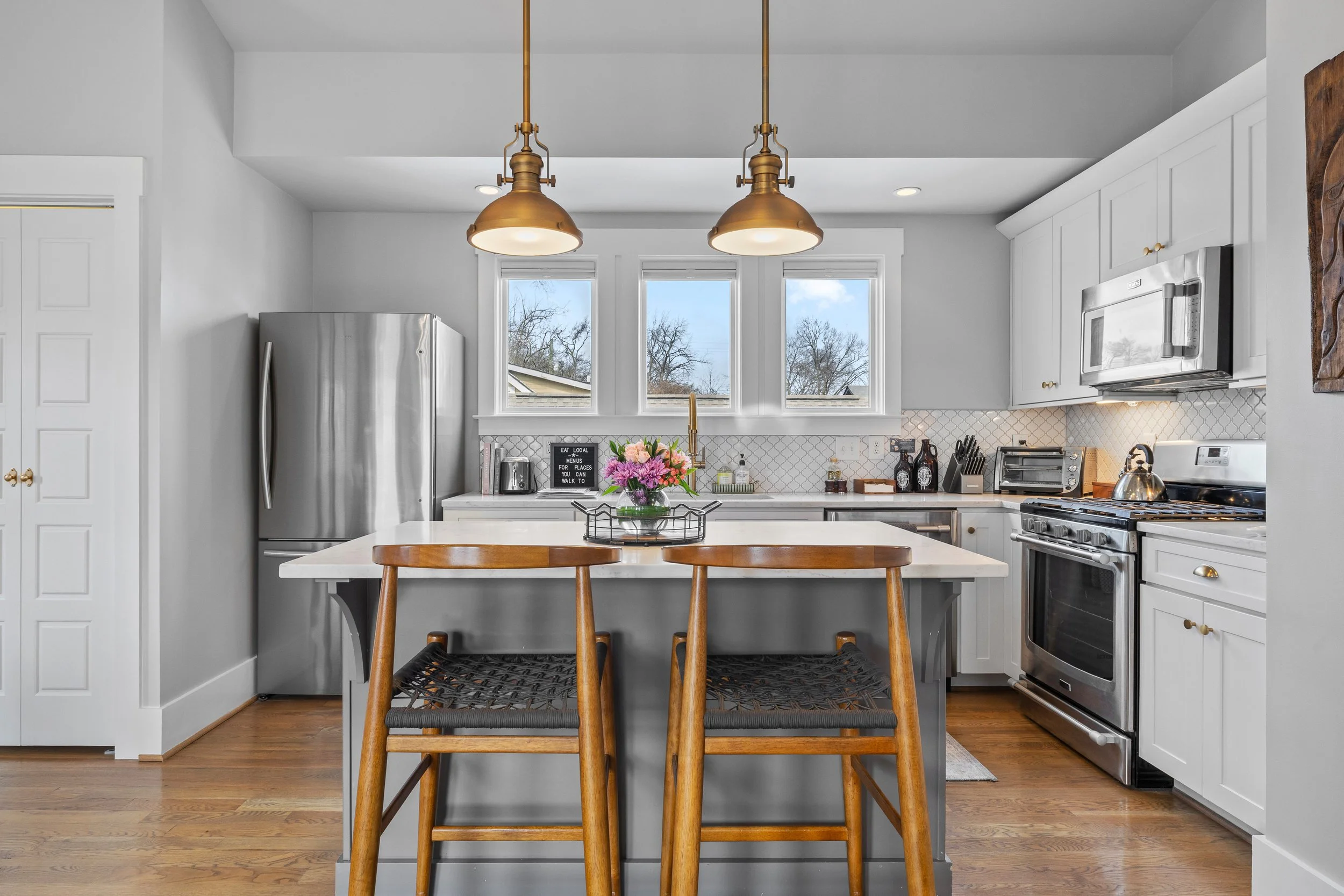 Modern white kitchen with stainless steel refrigerator, island with two wooden barstools, and pendant lights.