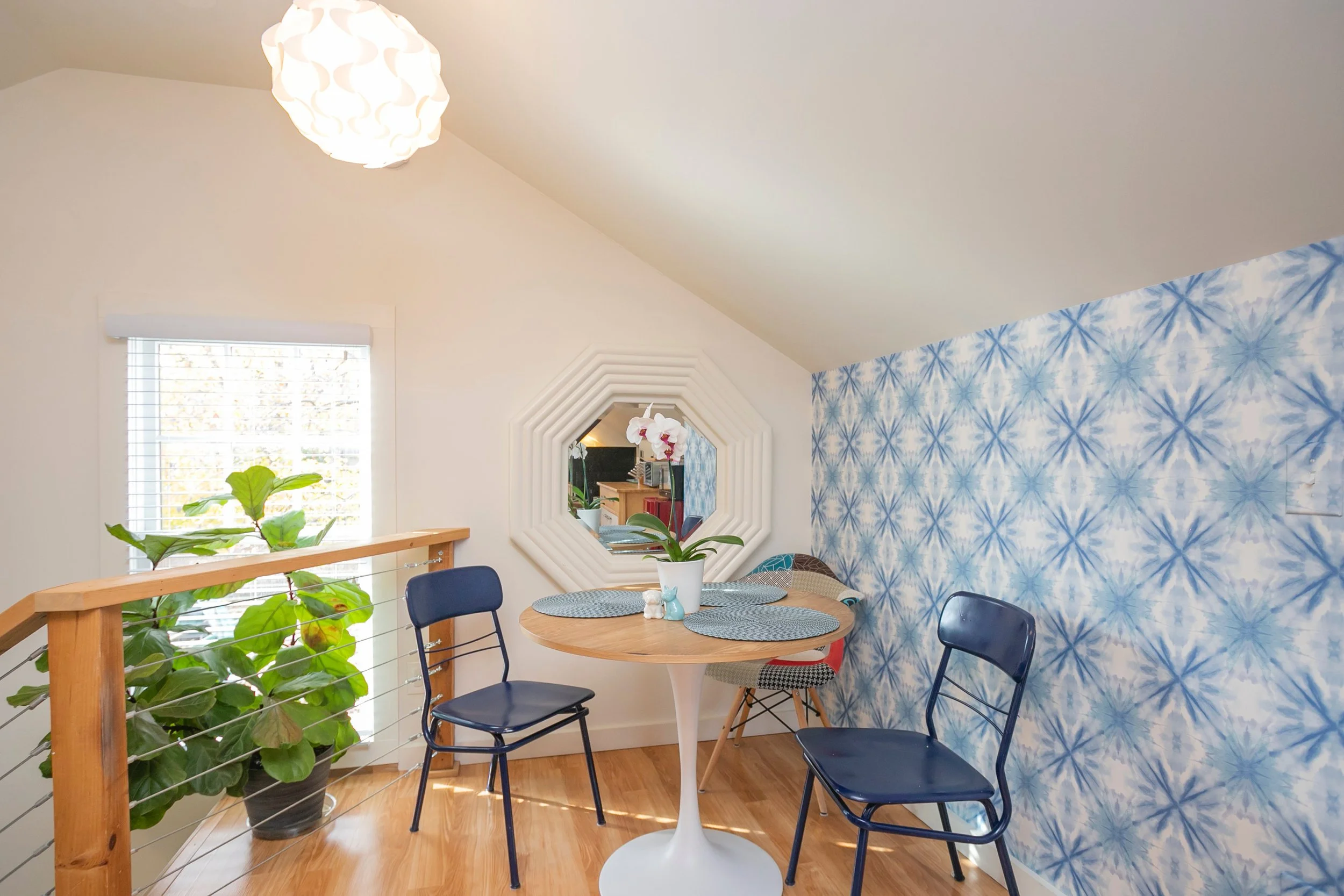 A cozy dining nook with a small round wooden table and four chairs, one with a colorful pattern. A large green plant is placed next to a window with white blinds. The wall behind the table features a blue and white tie-dye pattern, and a decorative white octagonal mirror hangs above the table. A white flower pot with pink orchids sits on the table, and a pendant light hangs from the ceiling.