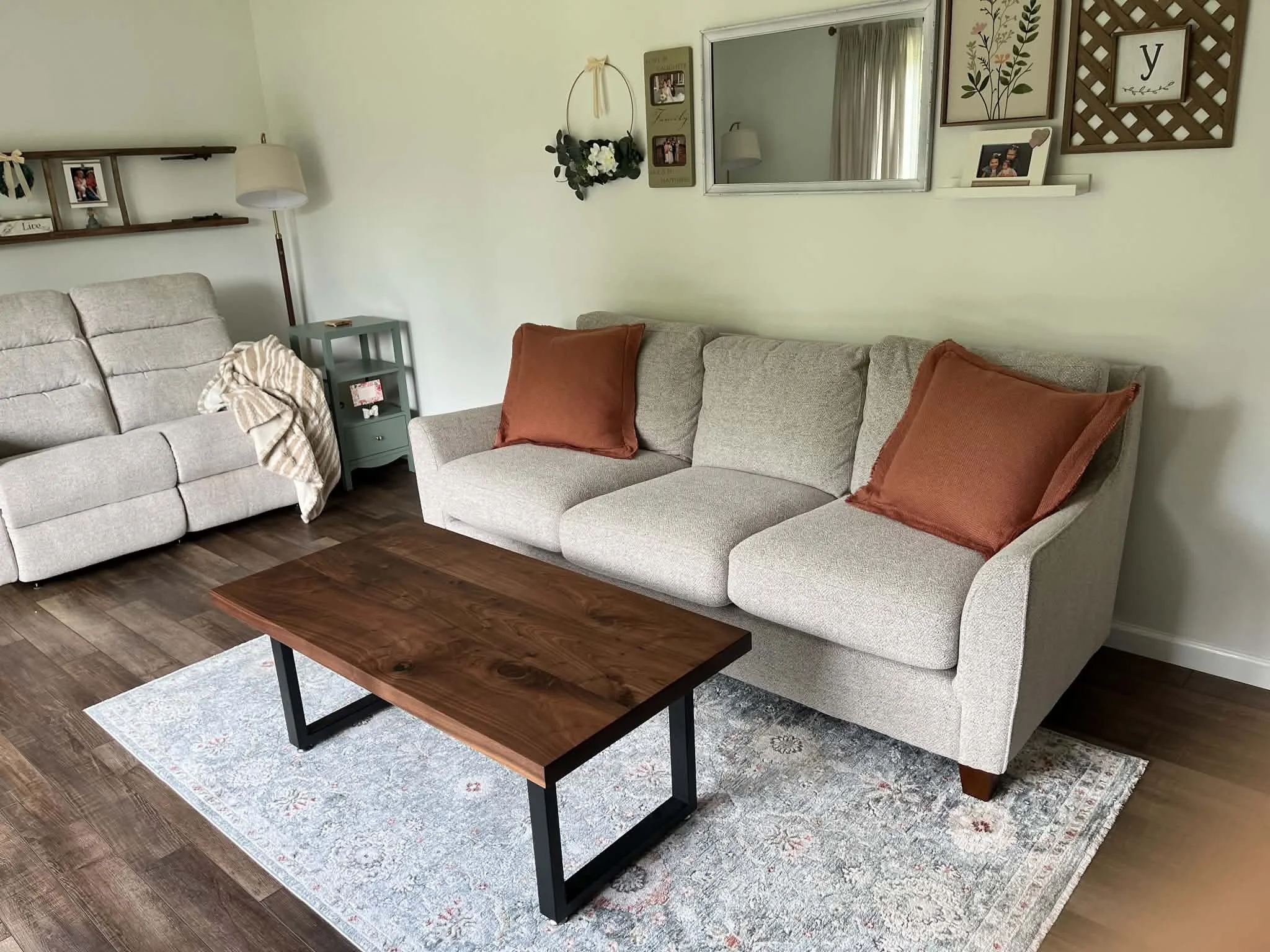 Living room with beige sectional sofa topped with orange pillows, a wooden coffee table, a gray side table, and a wall decorated with framed pictures, a mirror, and wall art.