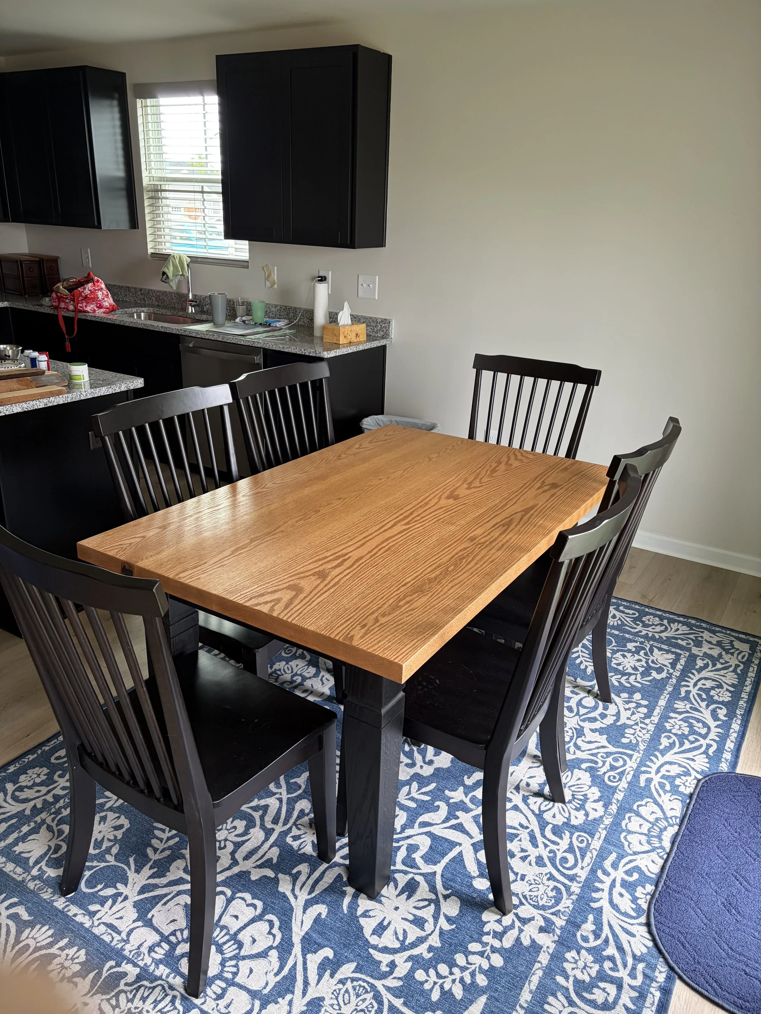 A dining table with six black chairs on a patterned blue and white rug, adjacent to a kitchen counter with black cabinets and a window with blinds.