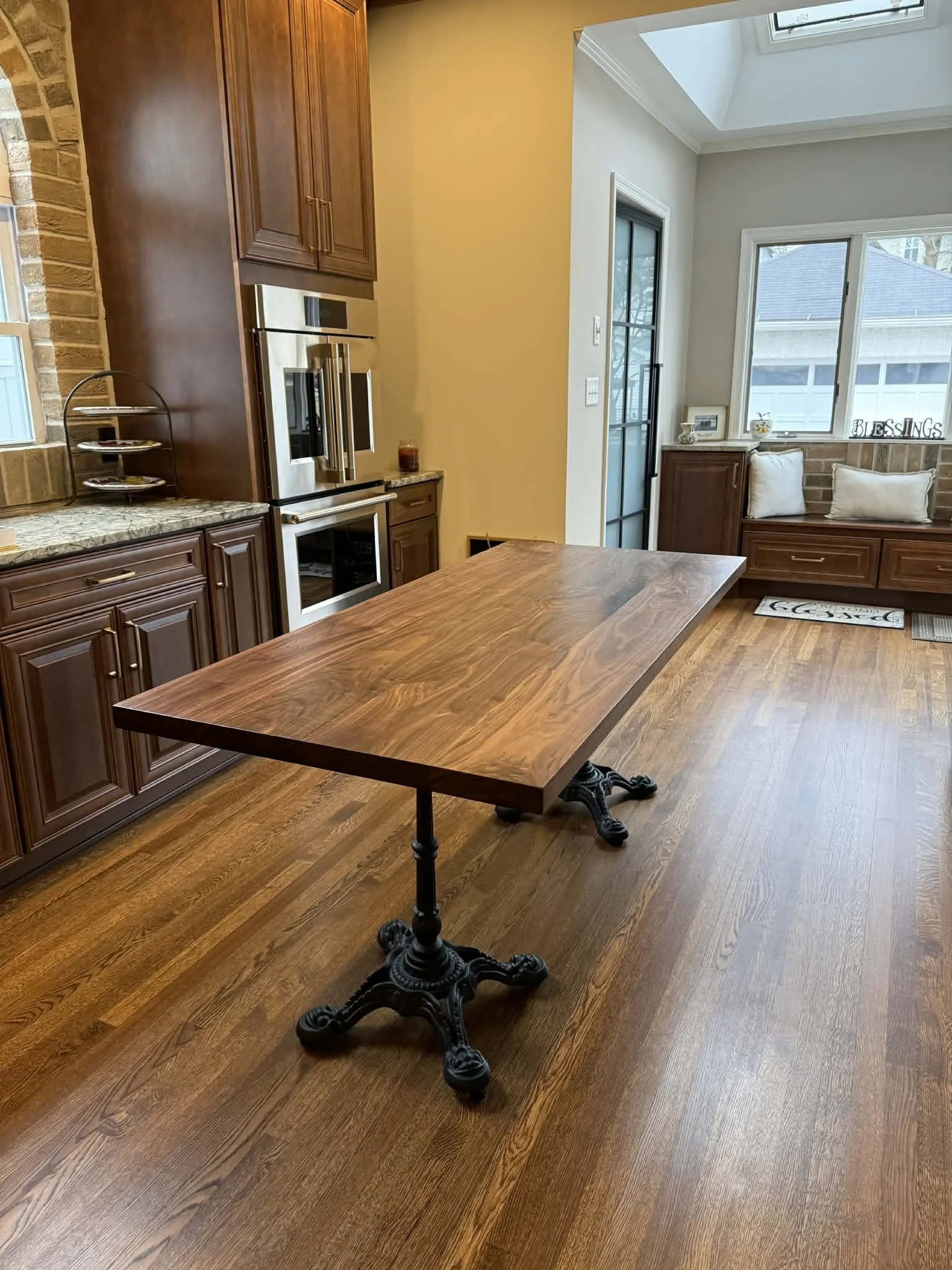 A wooden table in a kitchen with a decorative black metal base, surrounded by dark wood cabinets, a window seat with white pillows, and windows showing a neighboring house and trees.