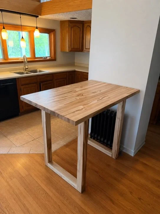 A wooden kitchen island table with a light natural finish, situated in a kitchen with wooden cabinets, a window with hanging pendant lights, and a hardwood and tile floor.