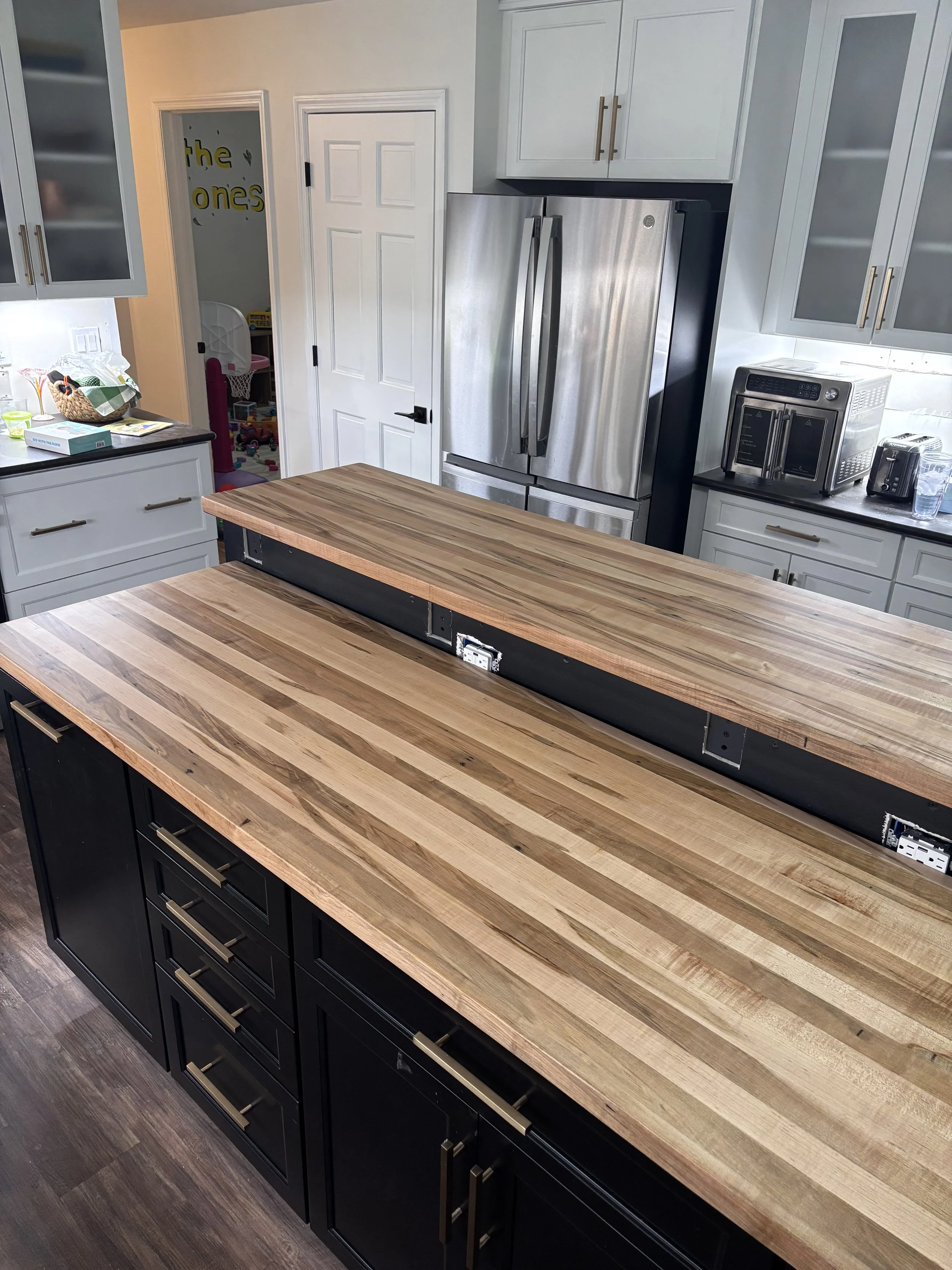 Kitchen with black cabinets and a wooden countertop, stainless steel refrigerator, microwave, toaster, and a partial view of a children's playroom through an open door.