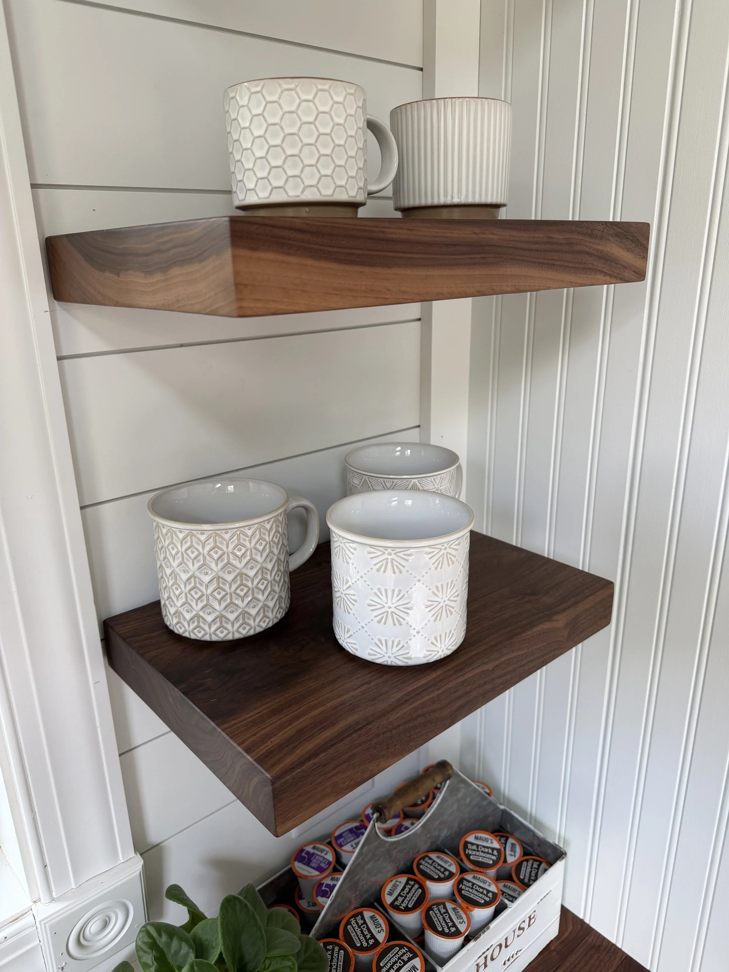 Two decorative ceramic mugs on a wooden shelf, with a white and beige geometric pattern, and a third plain beige mug on a lower shelf. A tray of black and orange CAULK TAPE containers is below, next to a green plant with heart-shaped leaves.