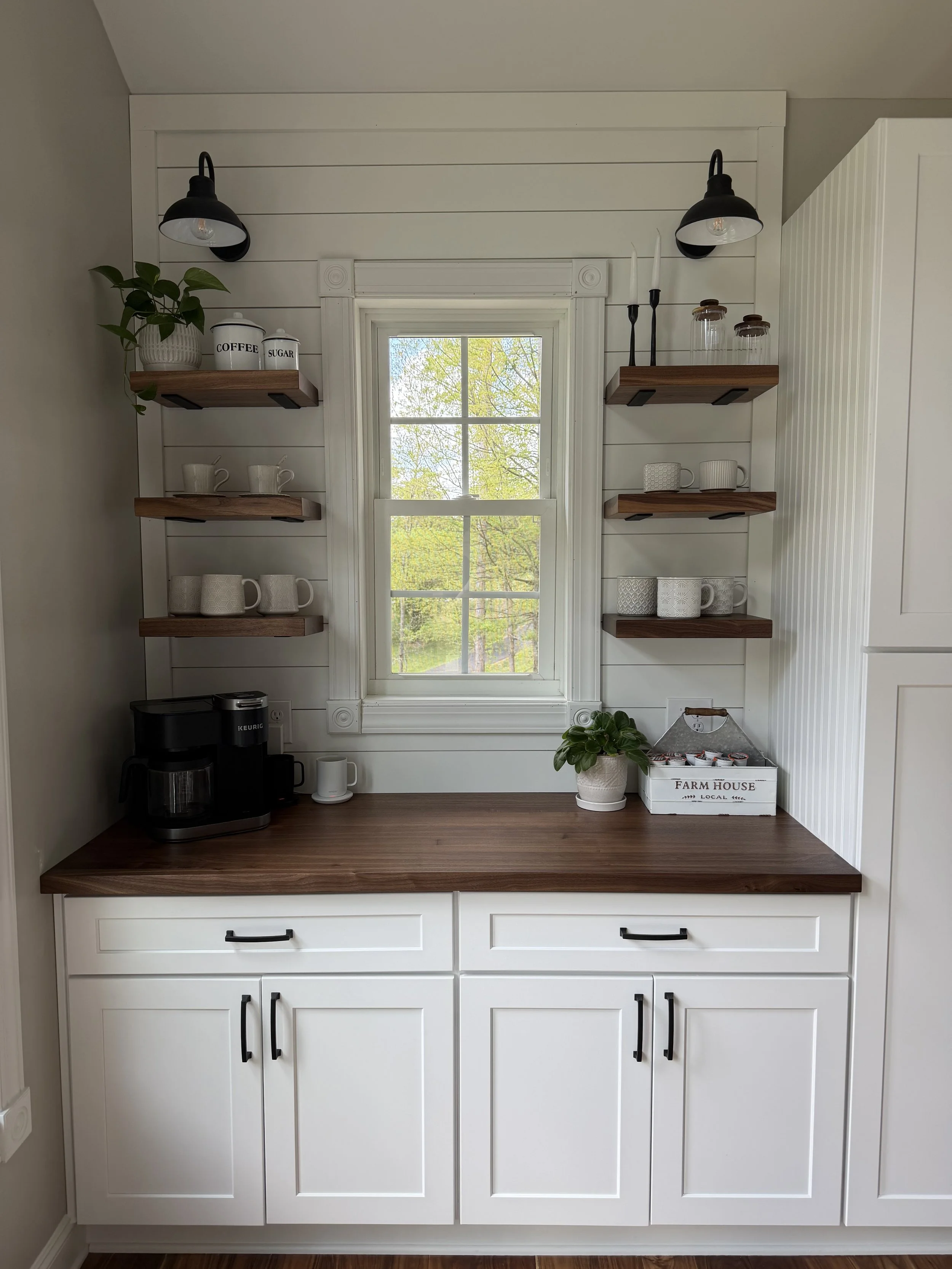 A kitchen corner with white cabinets, open shelves with cups, jars, and plants, a window with trees outside, and a wooden countertop with a coffee maker and cups.