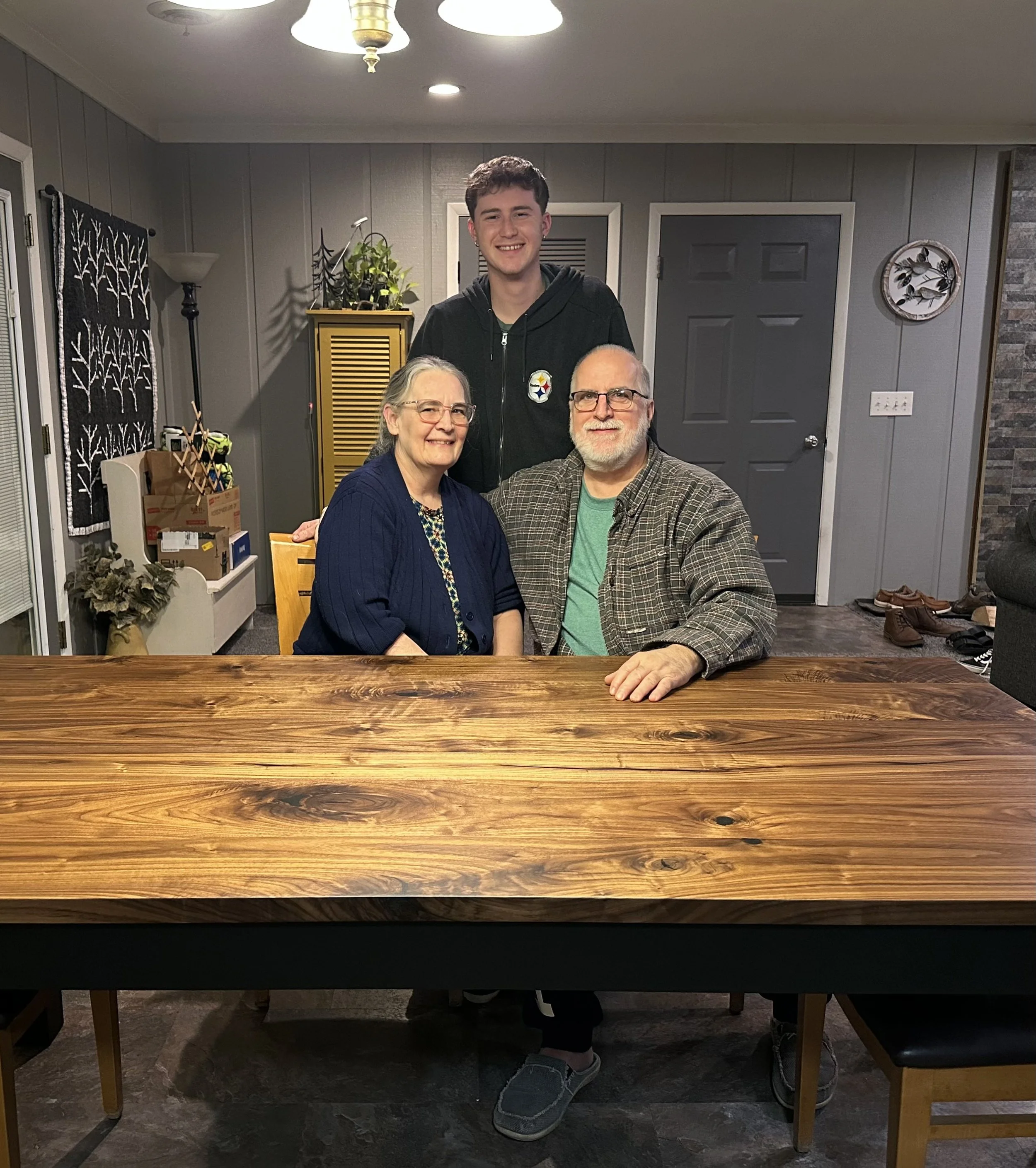 A family of three posing together in a dining room. The younger man is standing behind the seated older woman and man, all smiling at the camera. The dining room has gray paneled walls, a black door, a yellow cabinet with plants, and shoes on the flo