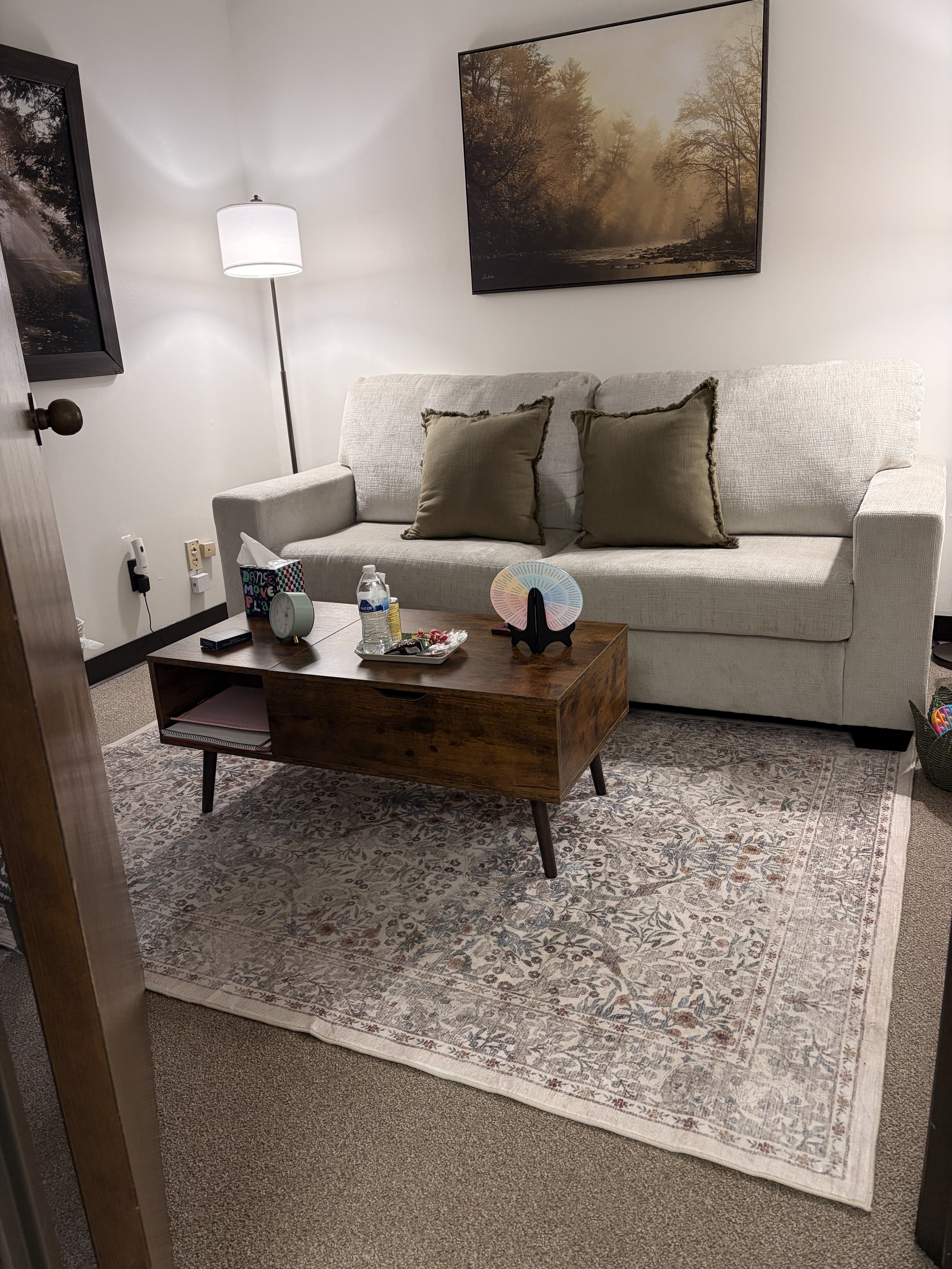 A cozy living room with a beige couch, decorative pillows, a wooden coffee table, two standing lamps, framed landscape artwork, an area rug, and a fireplace with a holiday garland on the wall.