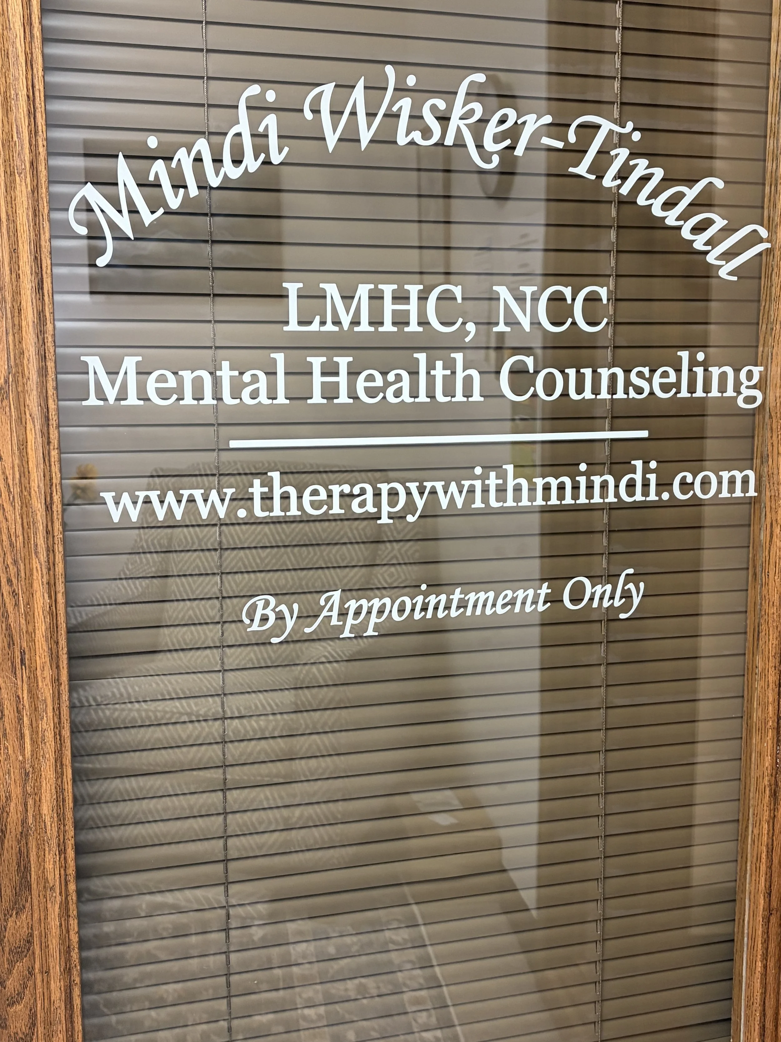 Wooden door with glass panels on either side, decorated with snowflake stickers and text indicating it leads to Mindi Wisker-Tindall's mental health counseling office. The door window has blinds, and the office interior is partially visible.
