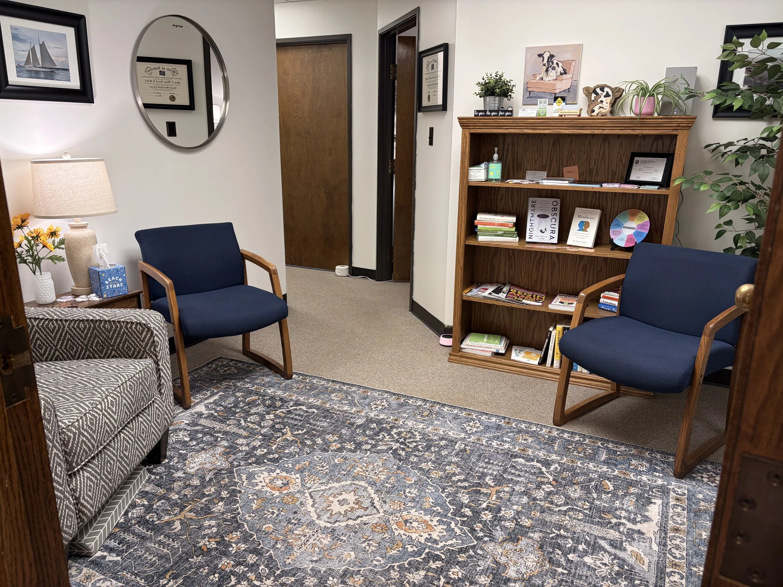 An office waiting room with two blue chairs, a patterned area rug, a wooden side table with a tissue box, a lamp, and a framed picture of a cow on a couch on the wall.