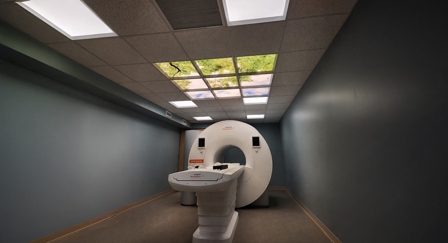 An empty medical imaging room with a Siemens Healthineers MRI machine and ceiling panels that resemble a sky with clouds and trees.
