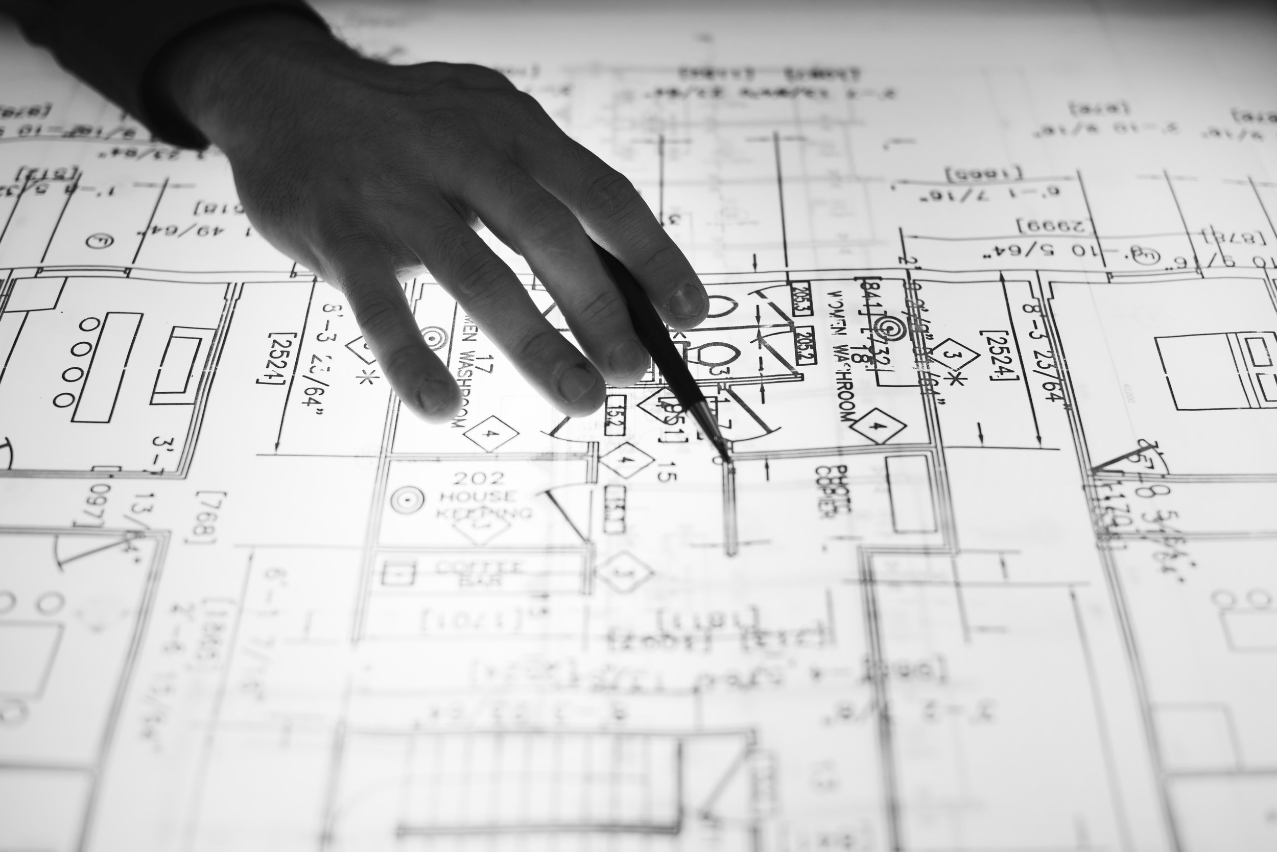 A hand holding a pen pointing to a technical blueprint or floor plan of a building.